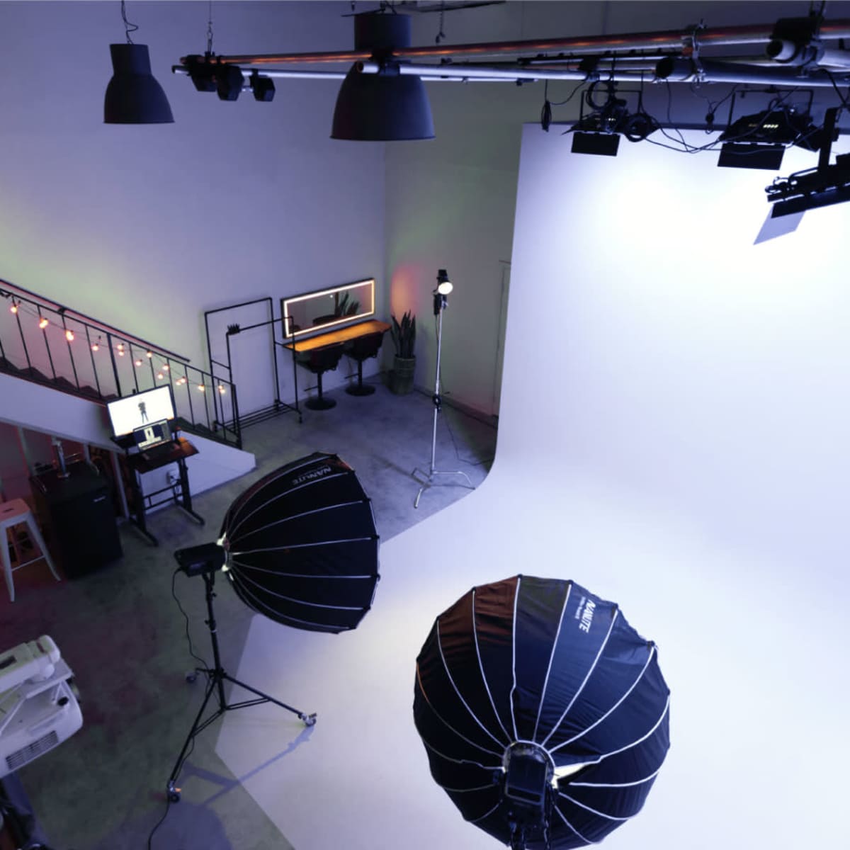 A photography studio features a large white backdrop, lighting equipment, and a workspace with a computer and seating on an upper level.