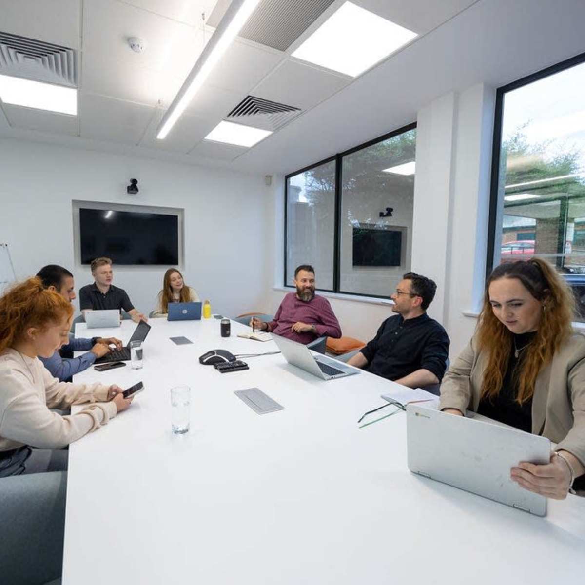 A group of six people is seated around a long table in a modern conference room, working on laptops and discussing ideas.