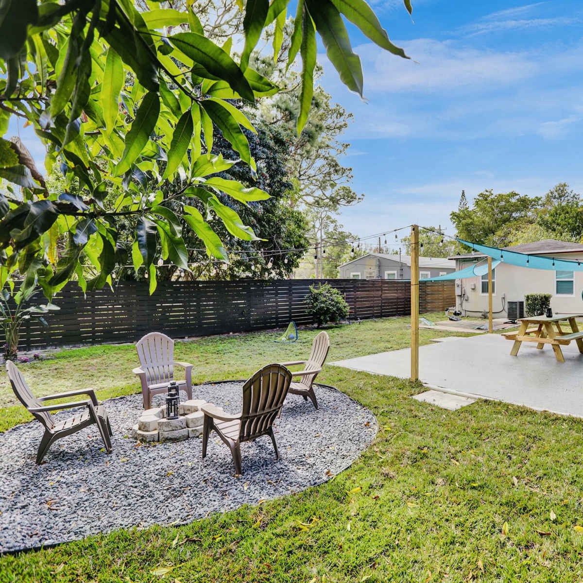A backyard with a fire pit surrounded by four chairs, a picnic table on a concrete patio, and a house in the background.