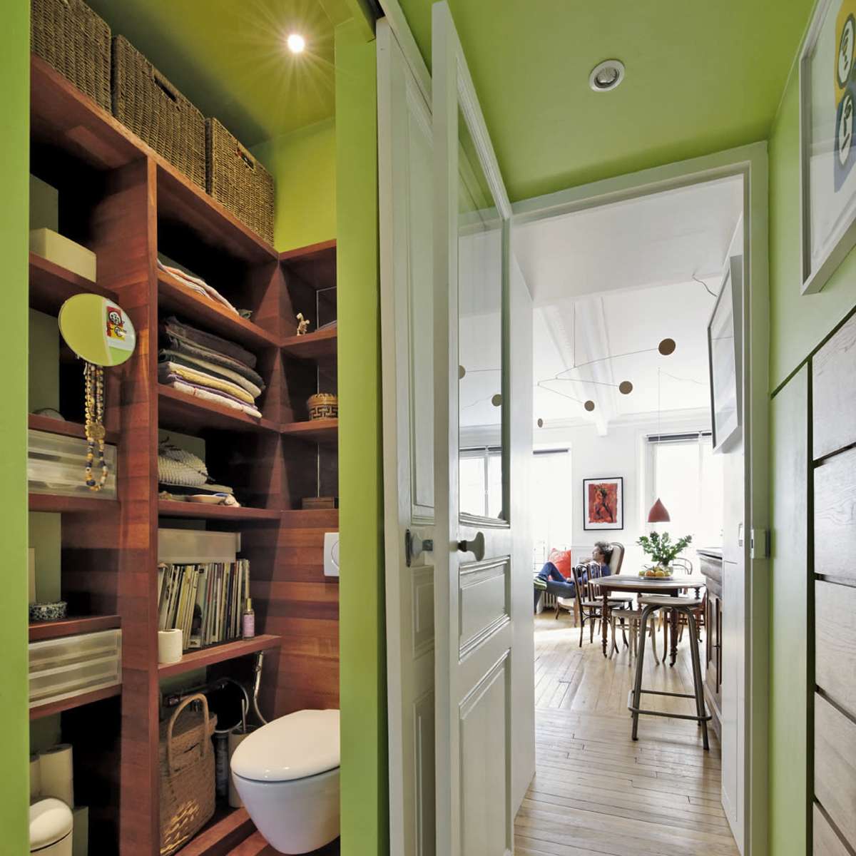 A narrow hallway leads to a bright green bathroom with shelves for storage and an open door that reveals a dining area in the background.