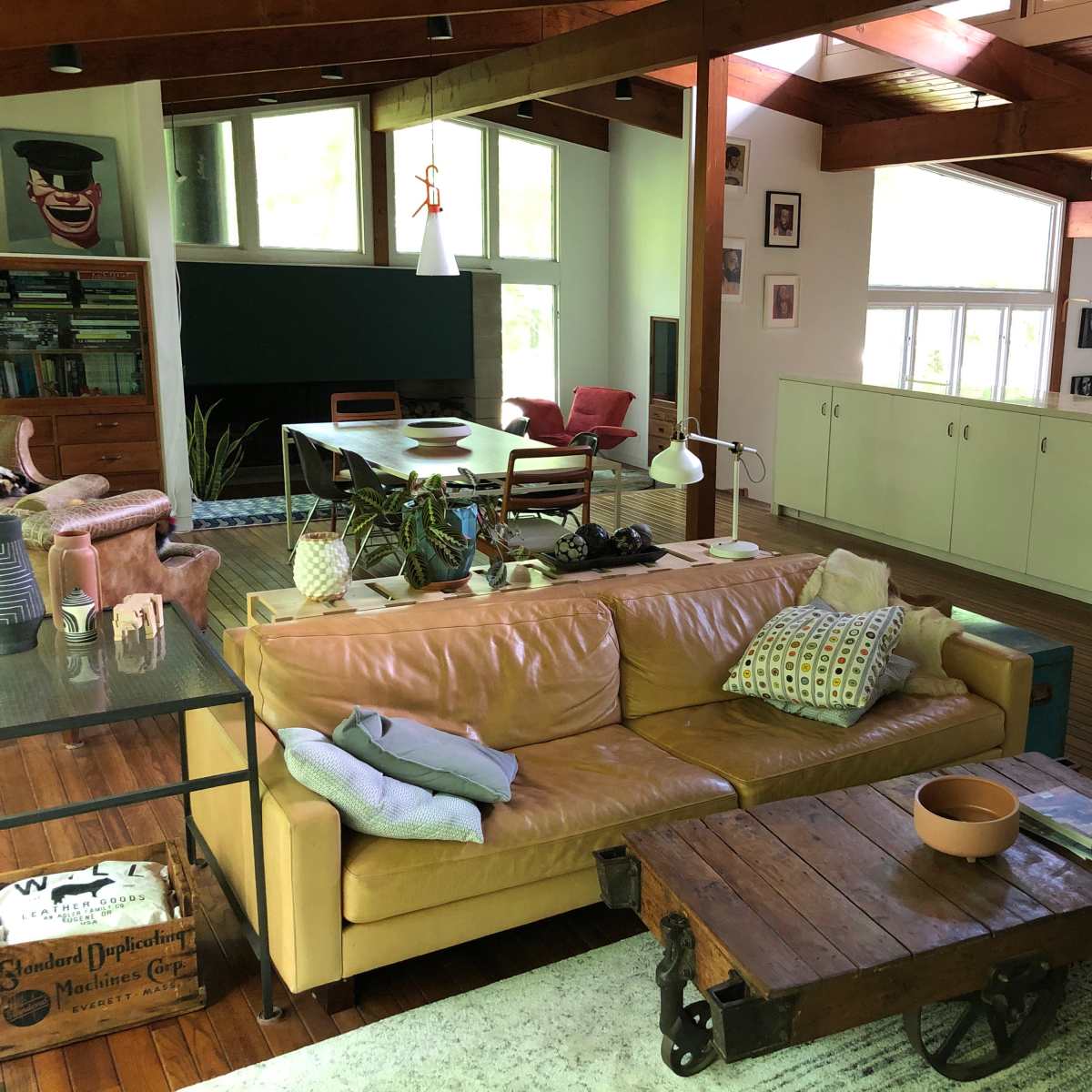 A spacious living area with a tan leather sofa, a wooden coffee table on wheels, a dining table in the background, and large windows letting in natural light.