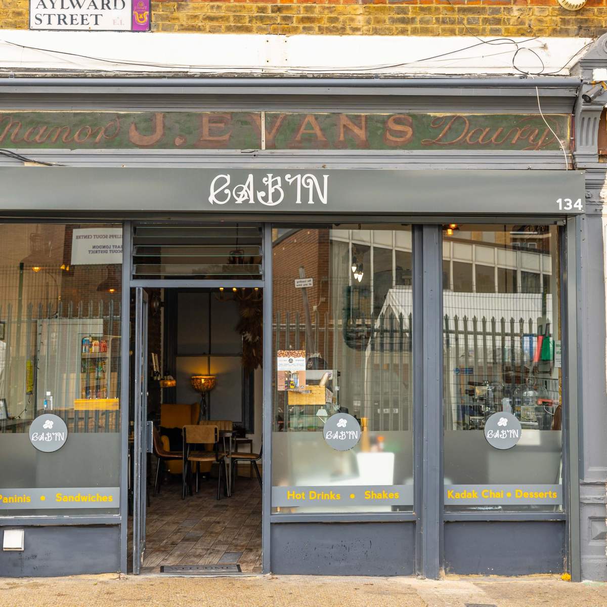 The storefront of a café named "CABIN," featuring large glass windows and a sign that includes the words "Paints & Sandwiches" and "Hot Drinks + Shakes + Kodak Eats & Desserts."