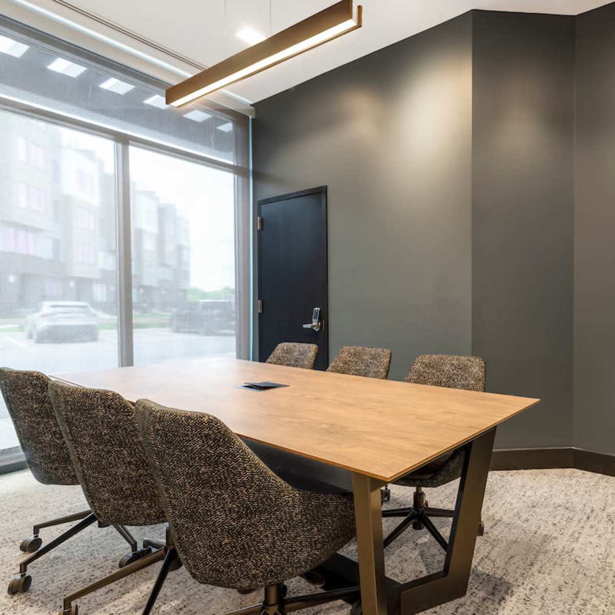 A conference room features a wooden table surrounded by four upholstered chairs and a green moss wall art on the gray wall.