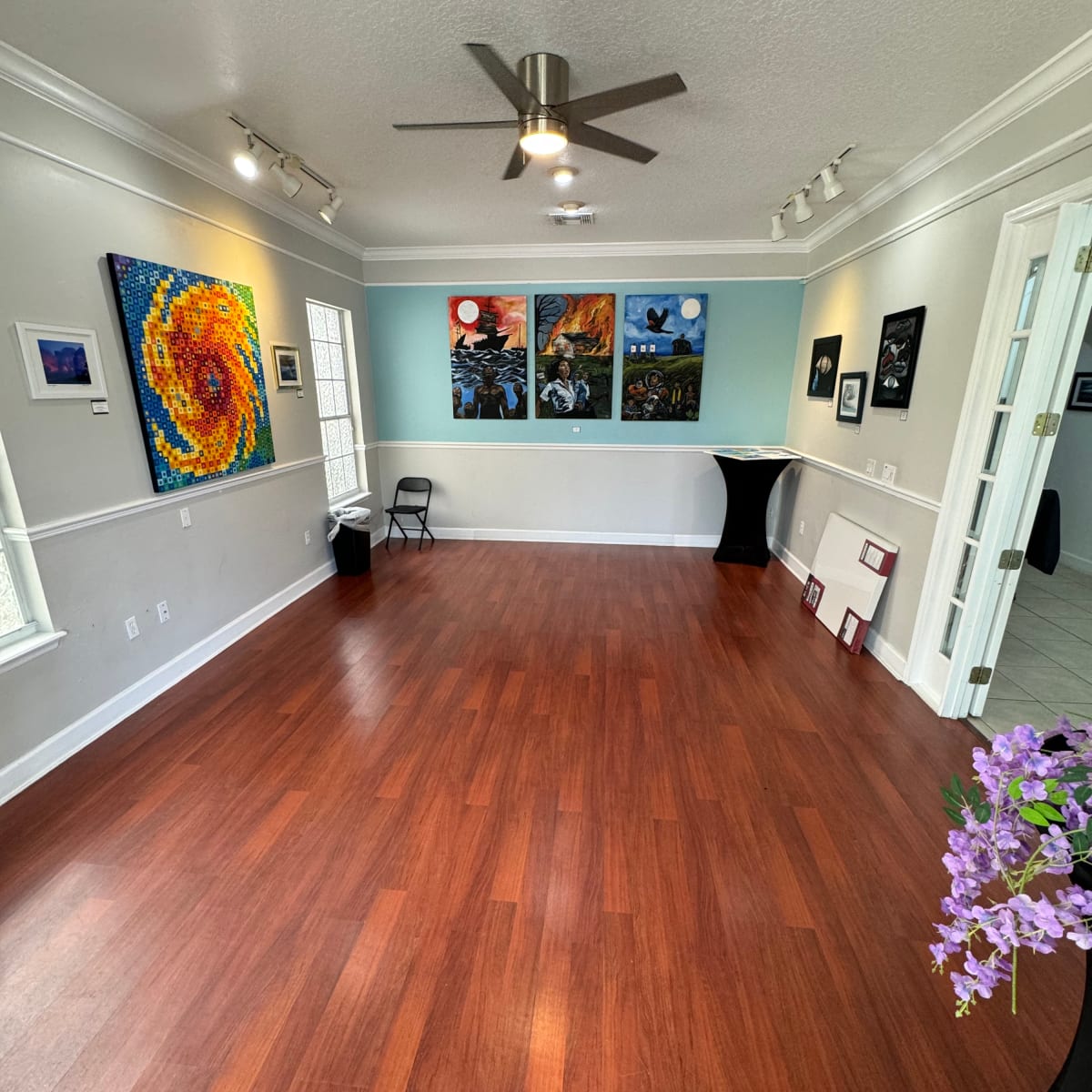 A brightly lit art gallery with wooden flooring, featuring colorful paintings on the walls and a small table in the corner.