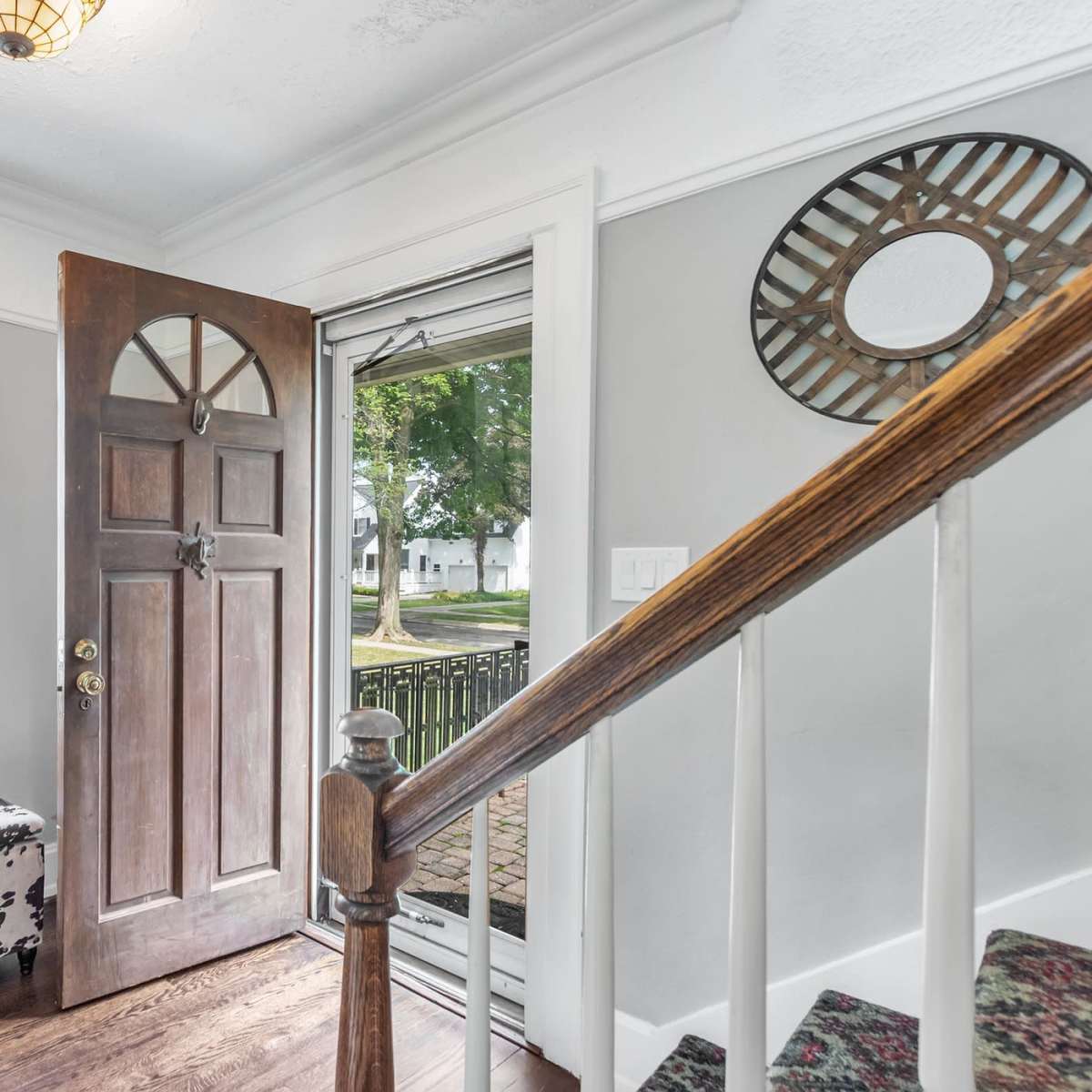 An entryway with a brown front door open to a view of a porch and exterior landscaping, along with a patterned bench and stairs leading to an upper level.