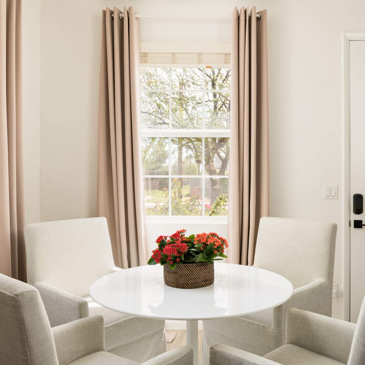 A small dining area features a round white table surrounded by four light gray chairs, with a potted plant in the center and large windows letting in natural light.