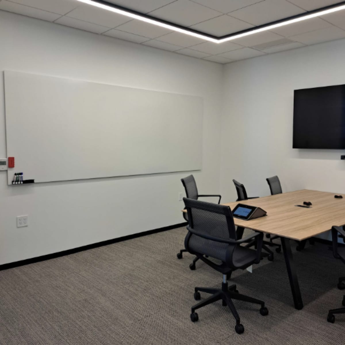 A modern conference room with a wooden table surrounded by five chairs, a large wall-mounted screen, and a whiteboard.