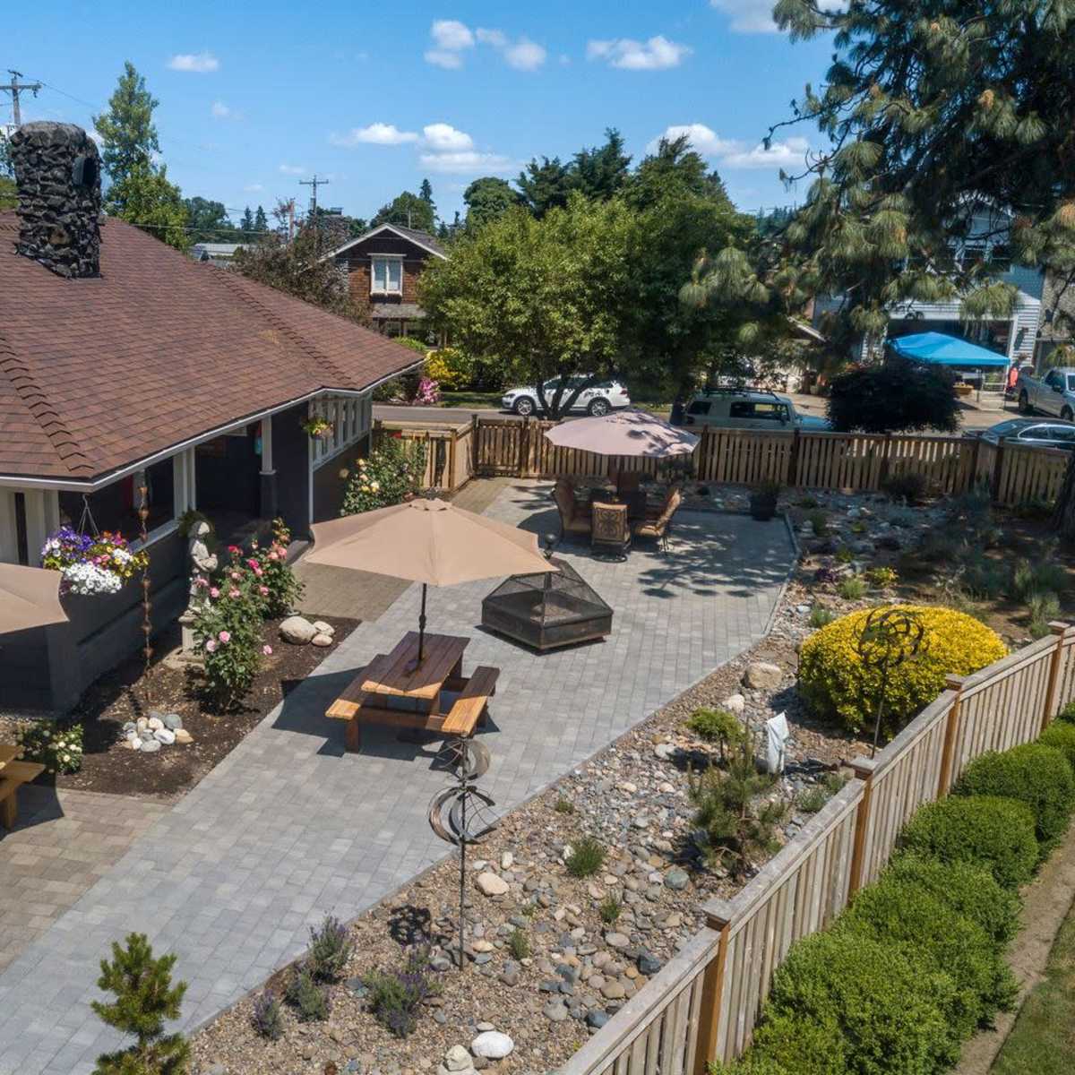 A backyard patio area with stone pathways, outdoor seating under umbrellas, and a landscaped garden surrounding a house.