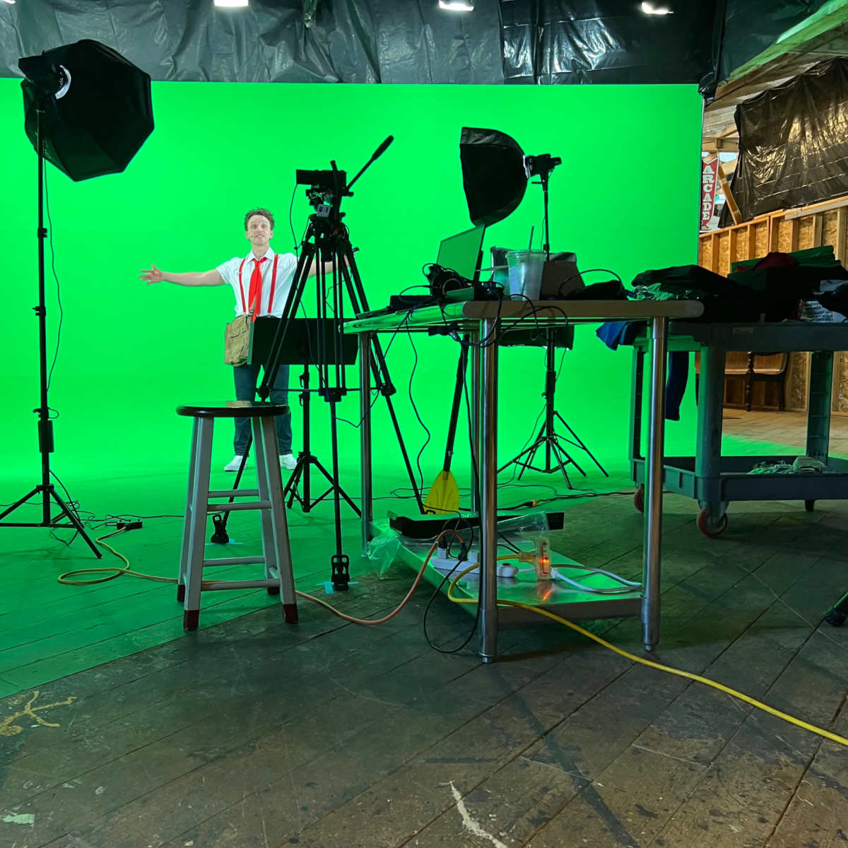 A person stands in front of a green screen with filming equipment and lighting set up around them in a studio.