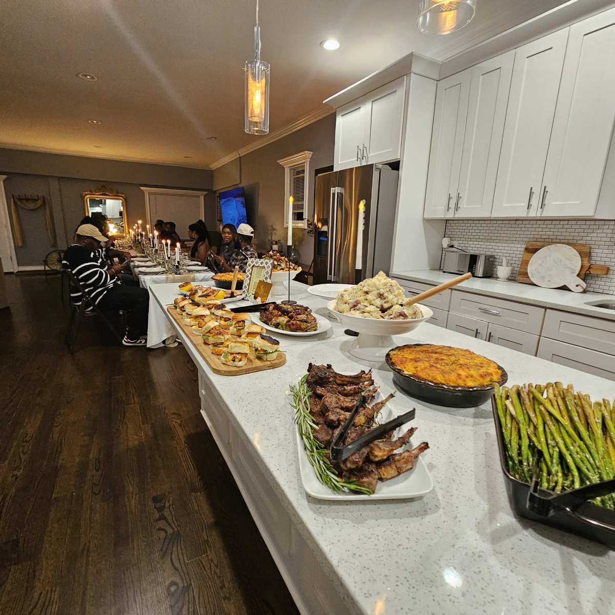 A beautifully arranged dining table features a variety of dishes, including roasted meat, appetizers, and sides, in a well-lit kitchen setting with guests seated nearby.