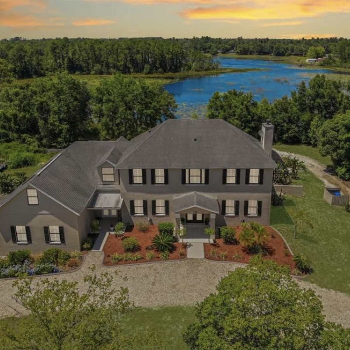 A large two-story house with a dark roof is set on a grassy landscape near a calm body of water, surrounded by trees and landscaping.