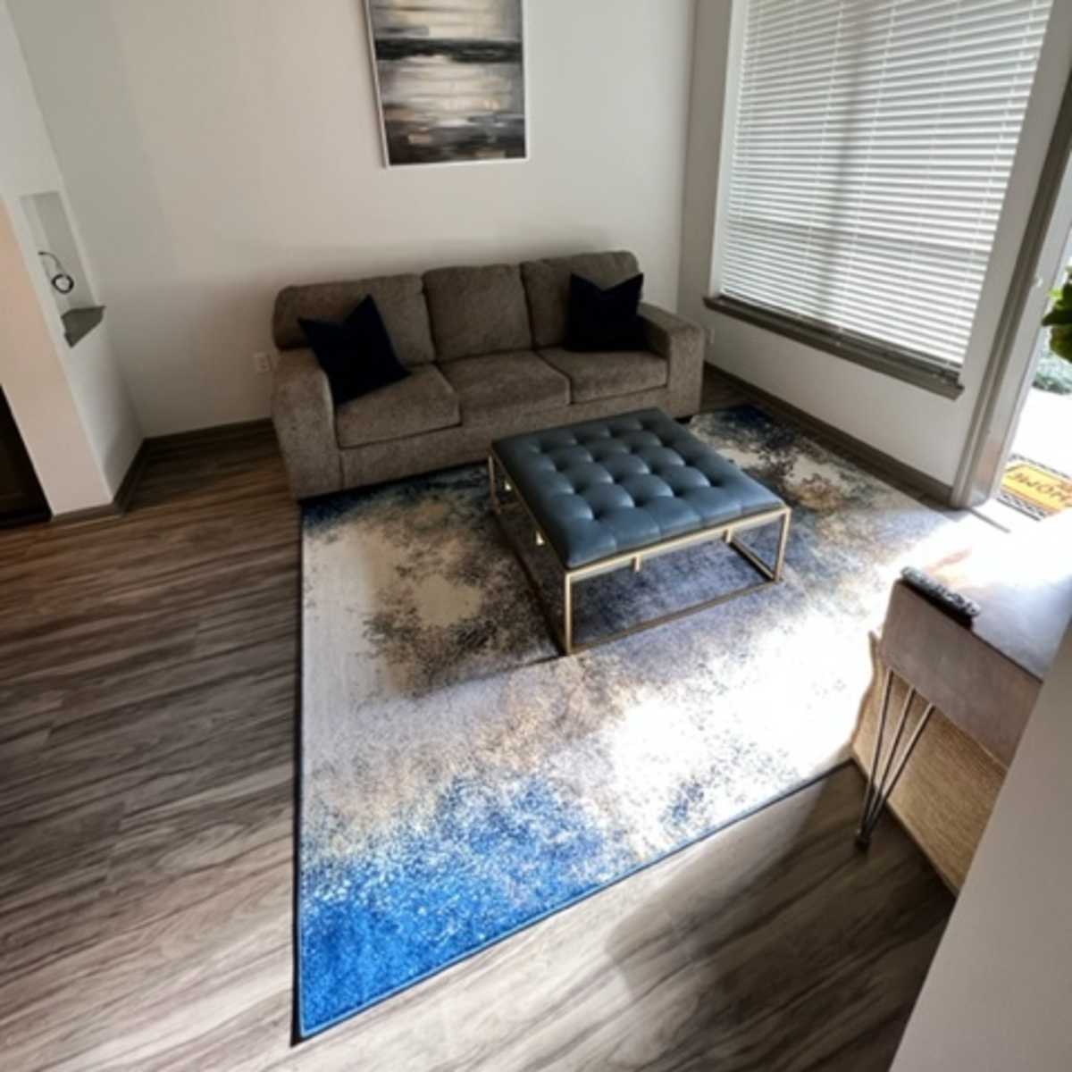 A modern living room features a gray sofa, a blue tufted coffee table, and a large area rug with a blue and beige design under a corner window with blinds.