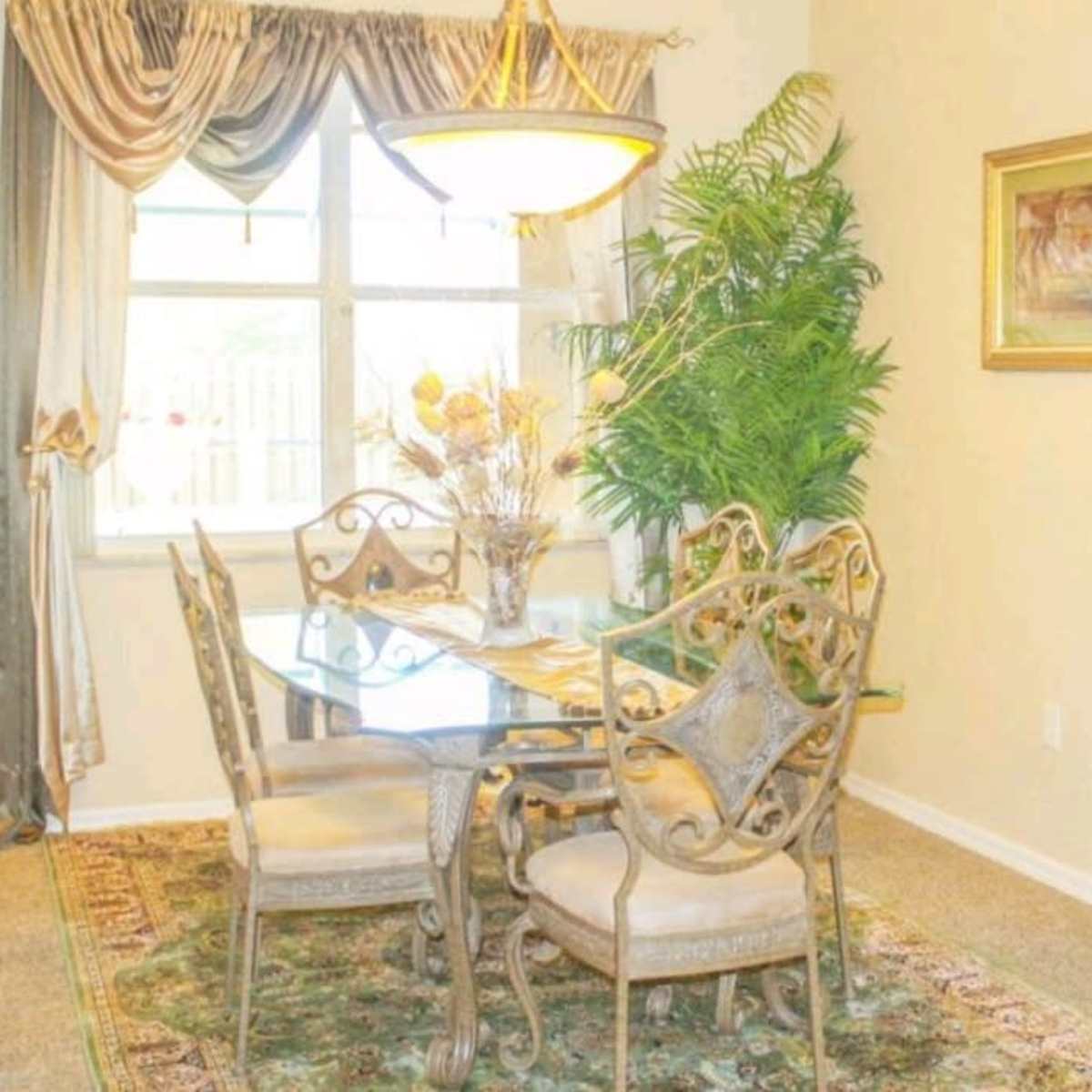 A dining area features a glass table surrounded by four ornate chairs, with a decorative plant and a painting accentuating the space.