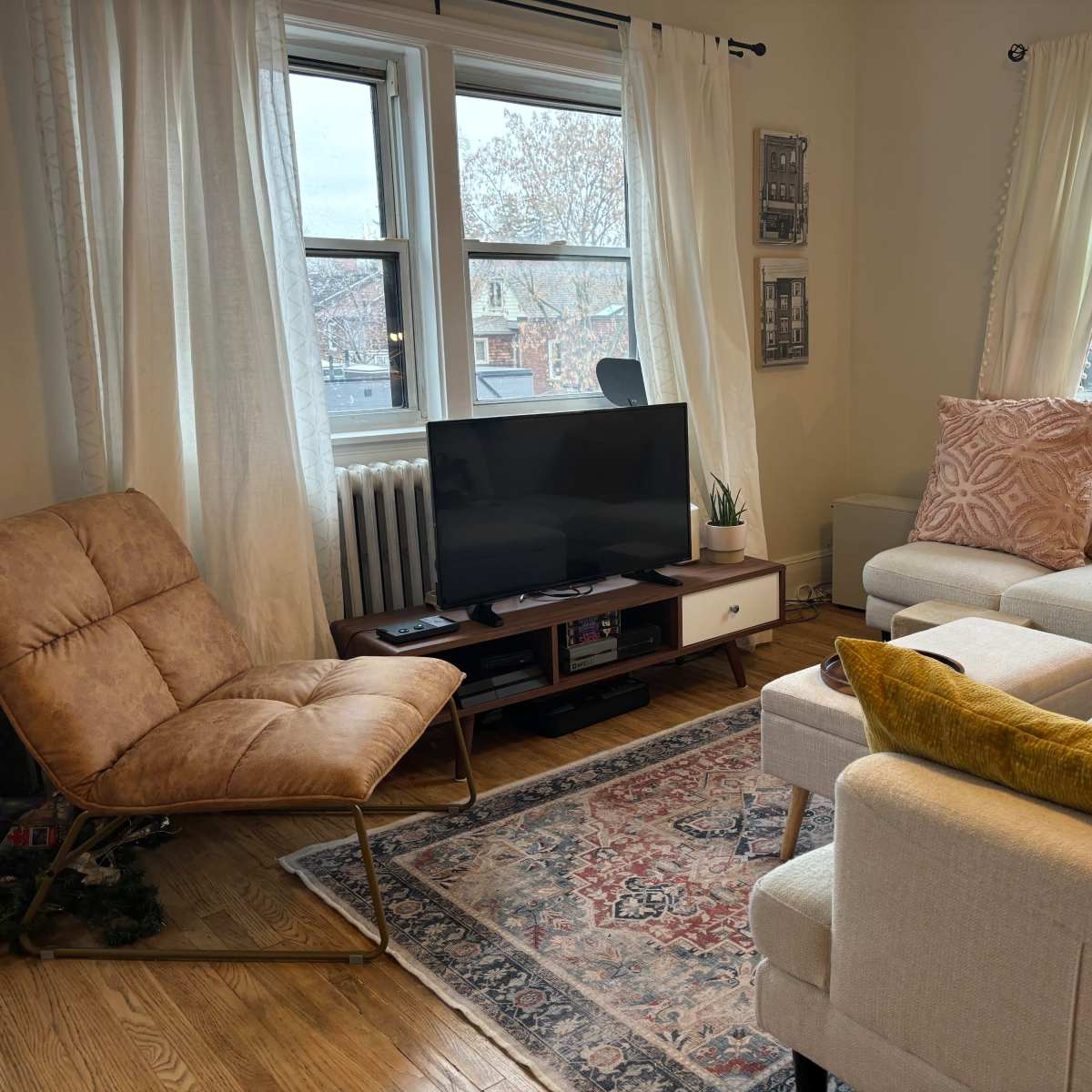 A cozy living room with a television on a stand, a light-colored sofa, a reclined chair, a decorative rug, and two large windows with sheer curtains.