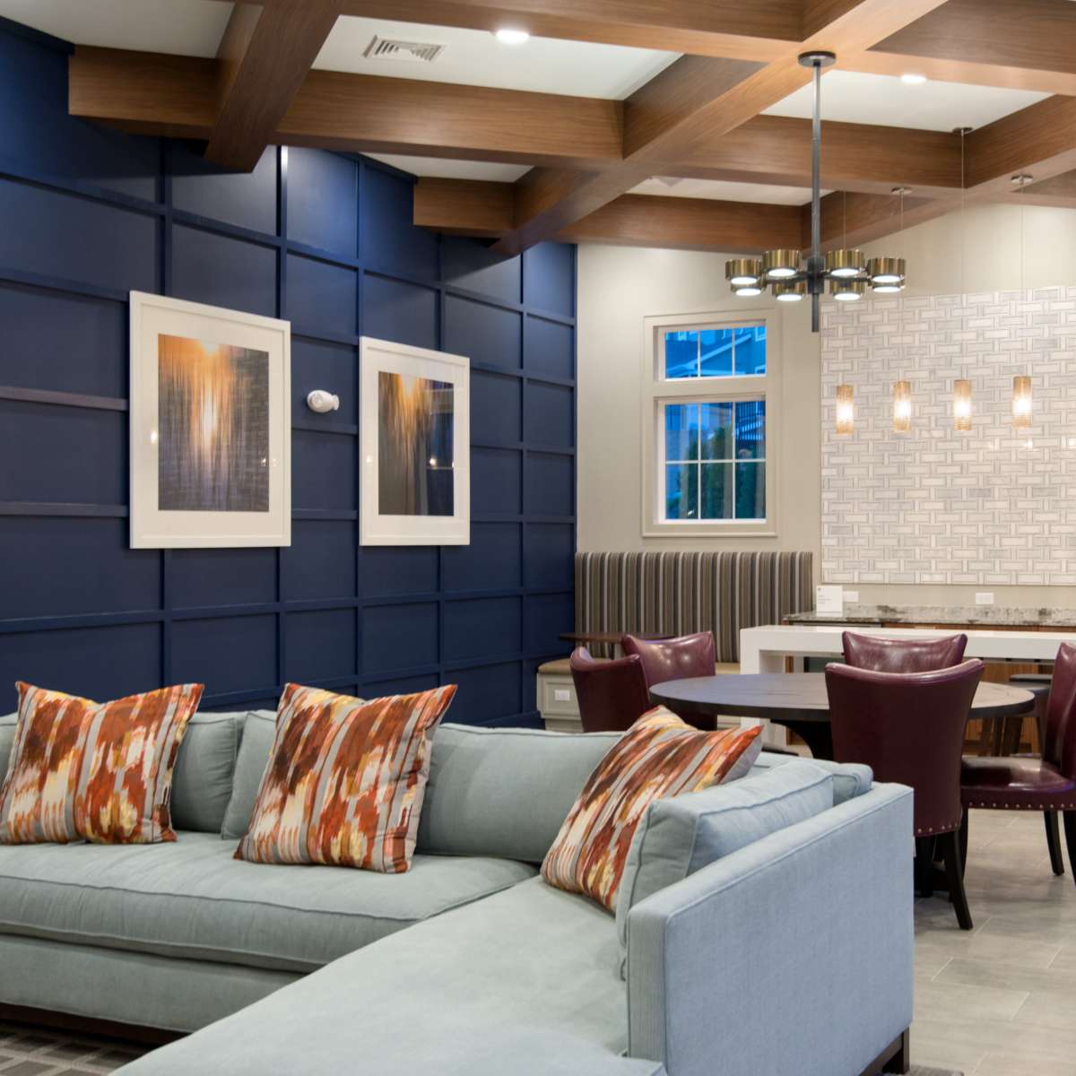 A modern lounge area featuring a light blue sectional sofa with decorative pillows, a dining area with a round table and burgundy chairs, and a kitchen space with white cabinetry and a tiled backsplash.