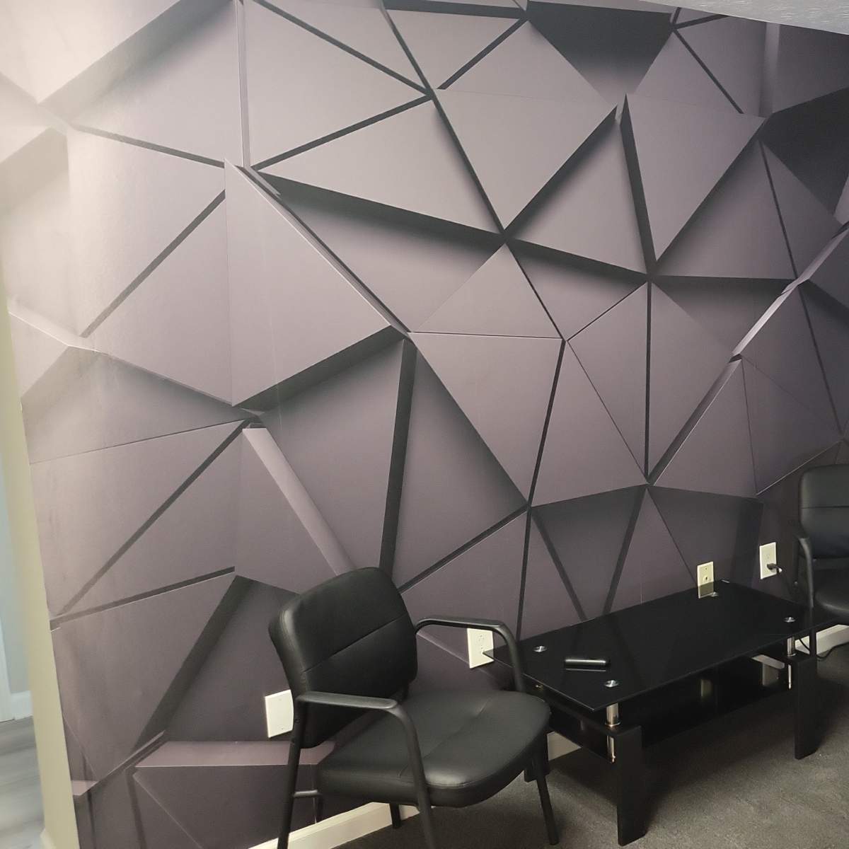 A small waiting area with two black chairs, a glass table, a geometric patterned wall, and a water cooler near an entrance.