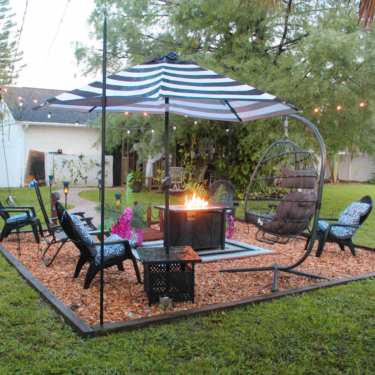A backyard seating area features a fire pit surrounded by chairs, a swinging chair, and a striped canopy, all set on gravel and grass.