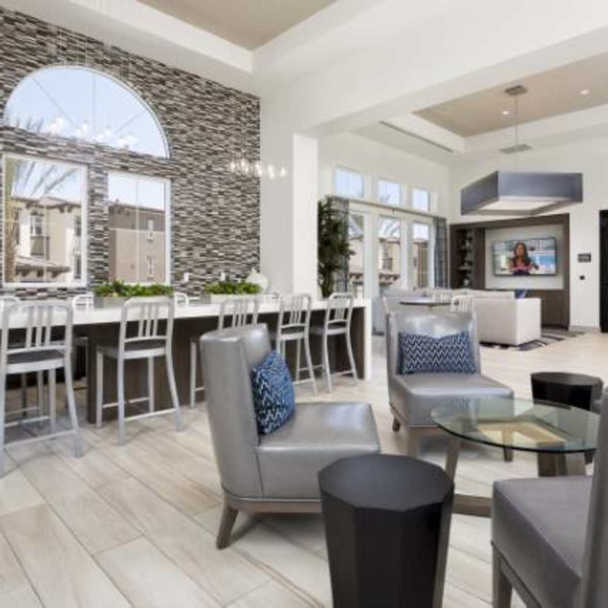 A modern indoor space featuring a kitchen bar with stools, a seating area with gray chairs, and large windows allowing natural light to enter.