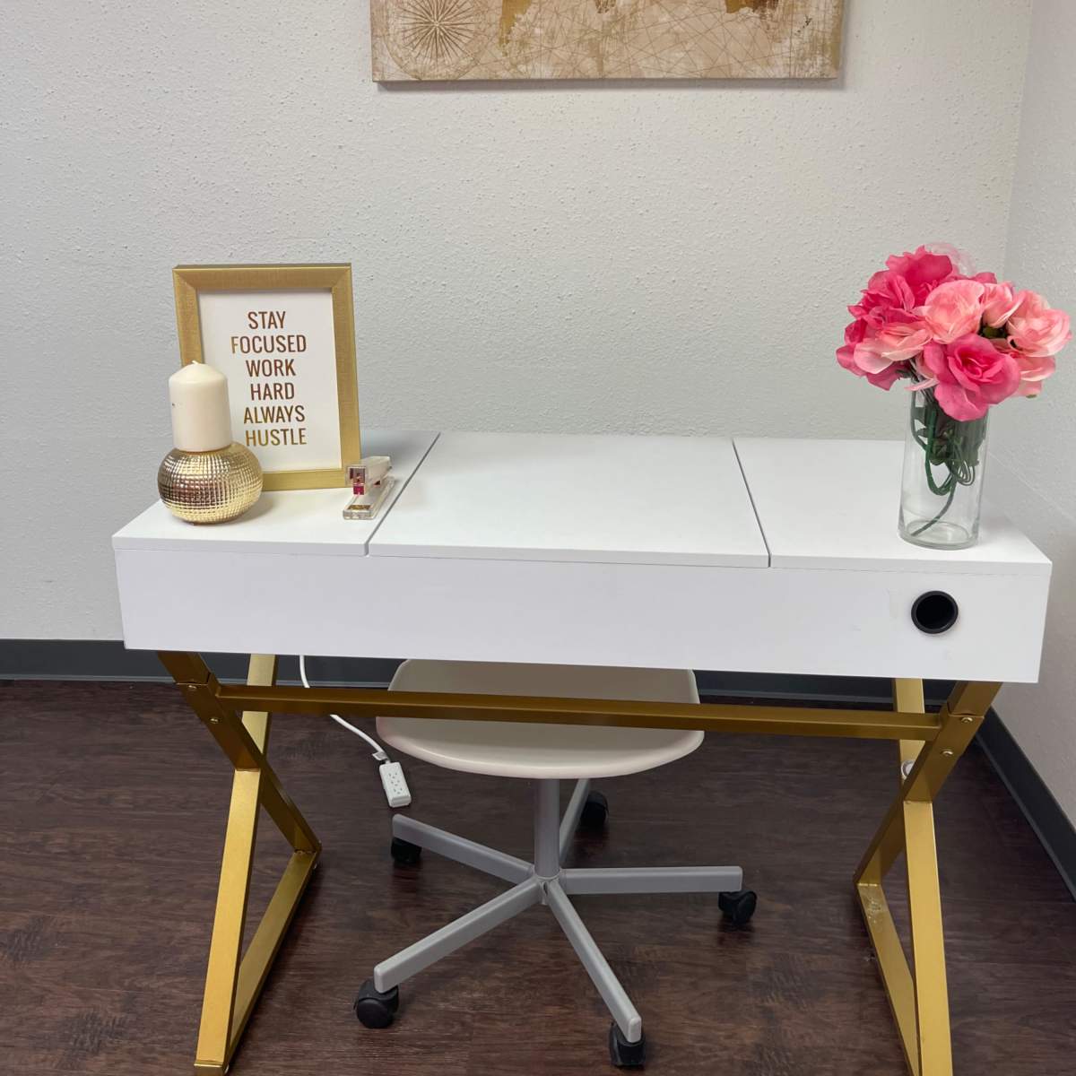 A white desk with gold legs is positioned against a wall, featuring a map art piece and decor items including a candle, a framed quote, and a vase of pink flowers.