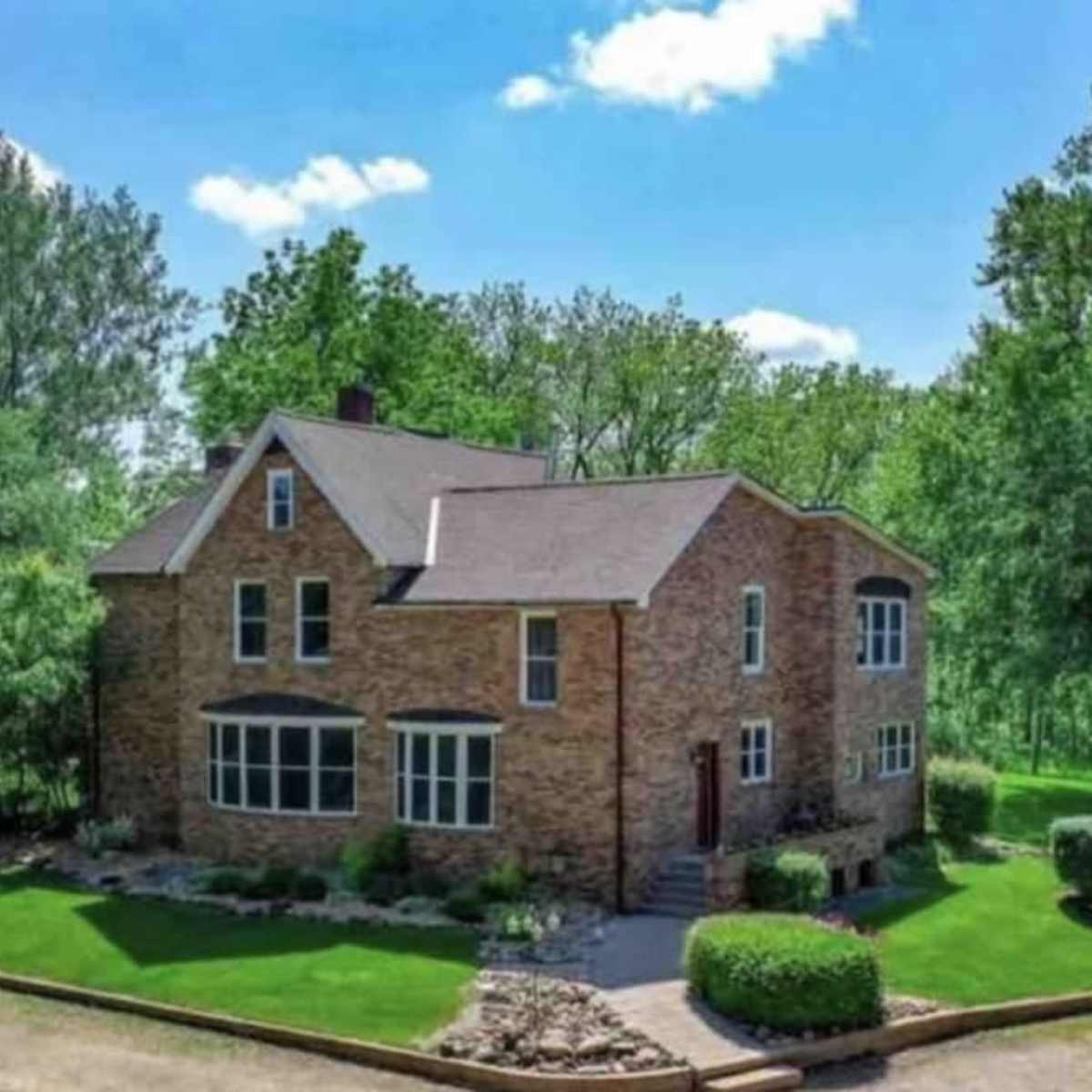 A large brick house with multiple windows is surrounded by grassy landscaping and trees under a partly cloudy sky.