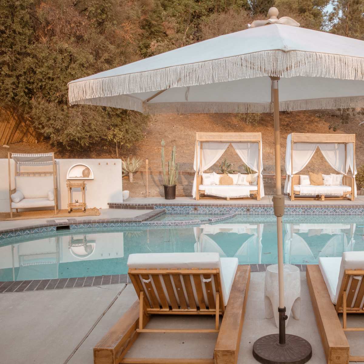 A pool area with two wooden loungers, an umbrella, and two canopied daybeds beside a landscaped hill.