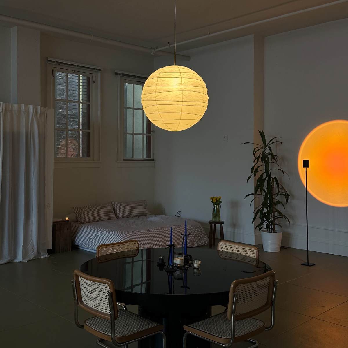 A round paper lantern hangs over a circular black table surrounded by four chairs in a softly lit room featuring a bed and a potted plant.