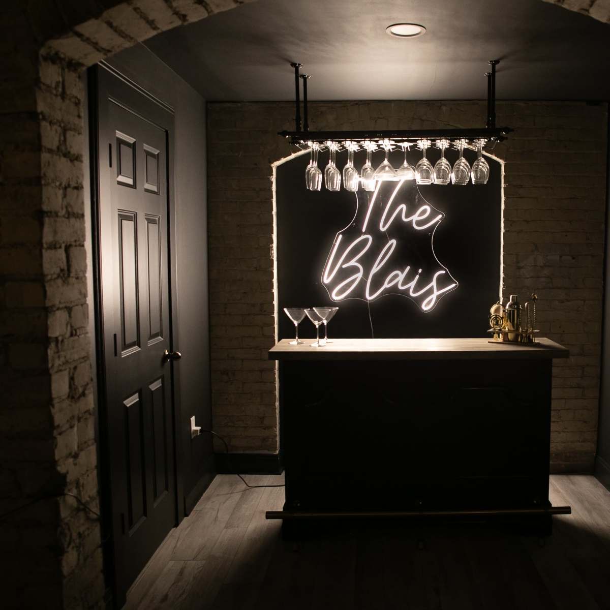 A dimly lit bar features a neon sign reading "The Blais" above a wooden counter lined with glasses.