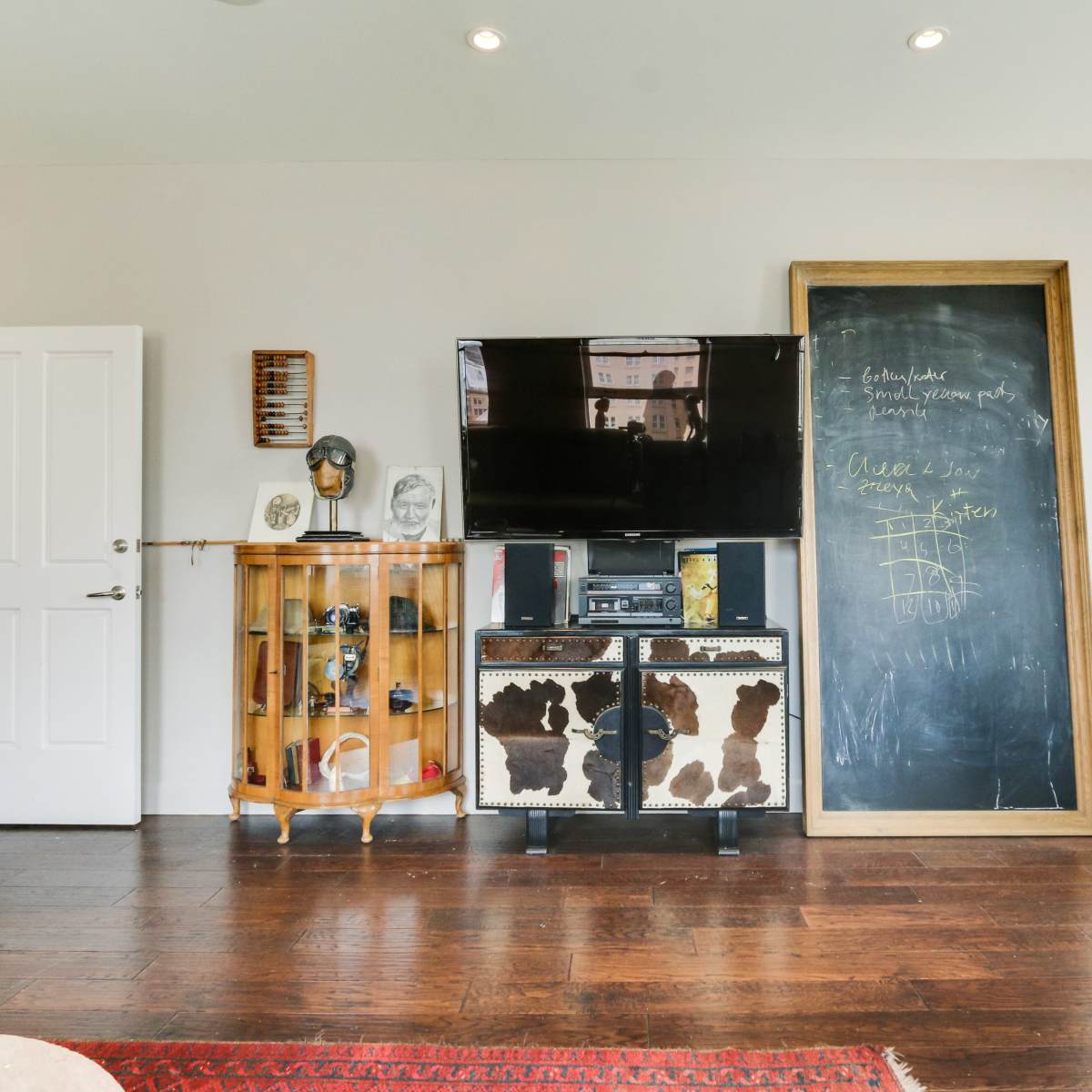A modern living space featuring a large television mounted on the wall, a wooden chalkboard, and a vintage cabinet in front of dark wood flooring.