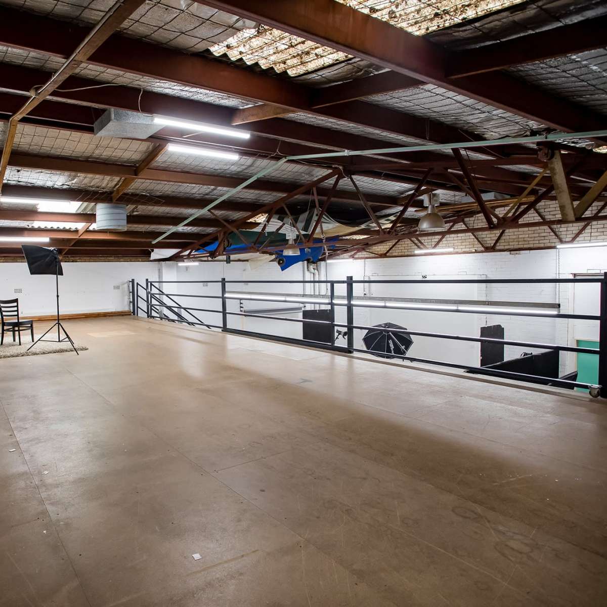 A large, empty indoor space with high ceilings, metal beams, and a wooden floor, featuring a chair and lighting equipment set up for a photo shoot.
