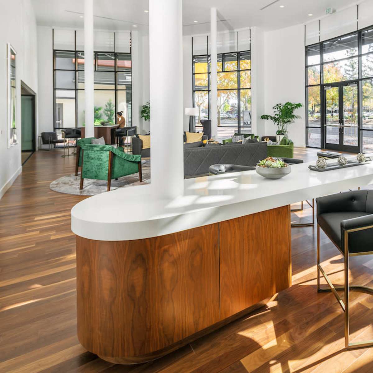 A spacious, modern lounge area features a large white countertop bar with black stools, surrounded by tall windows that let in natural light and views of greenery outside.