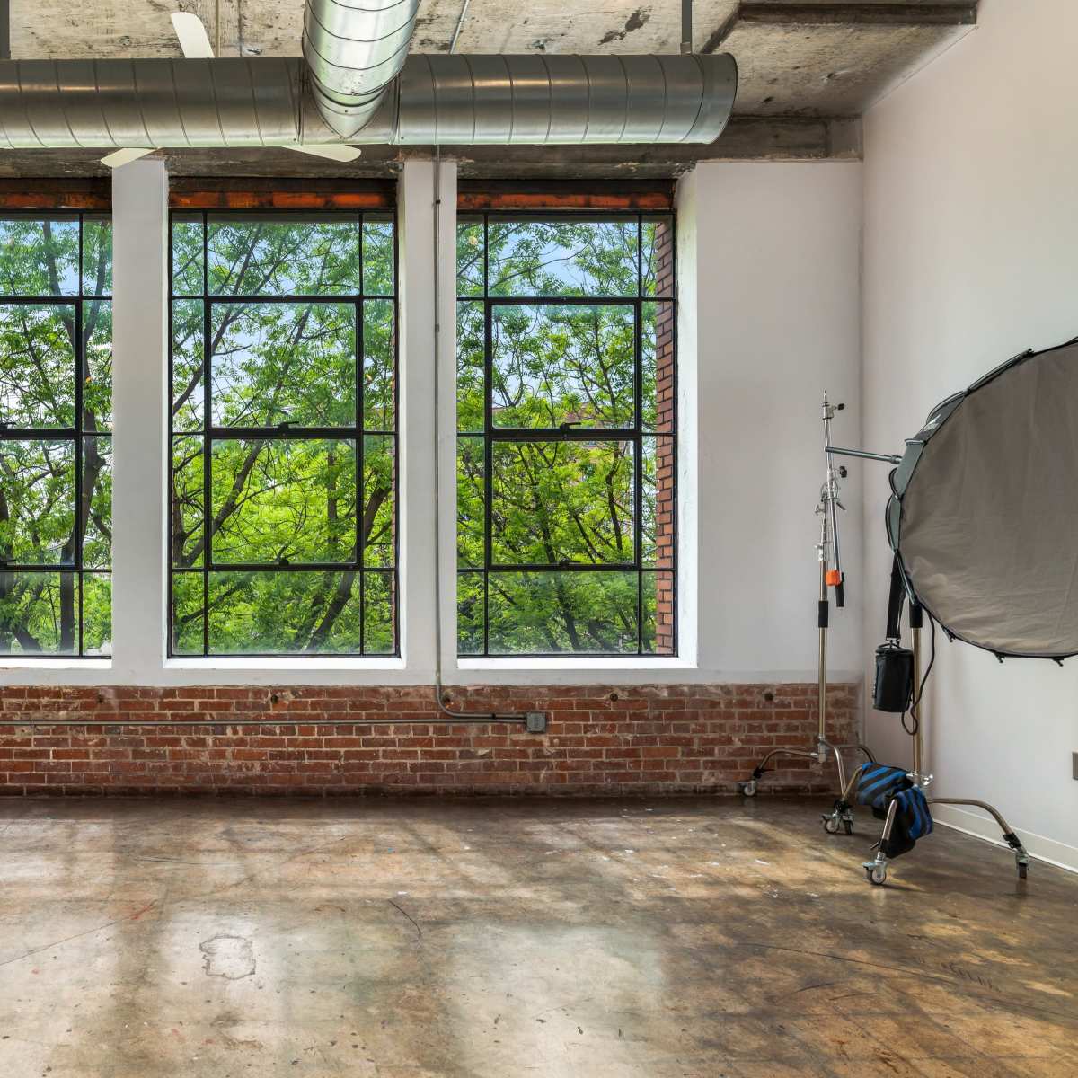 A spacious room with large windows overlooking green trees, featuring exposed brick walls and concrete flooring.