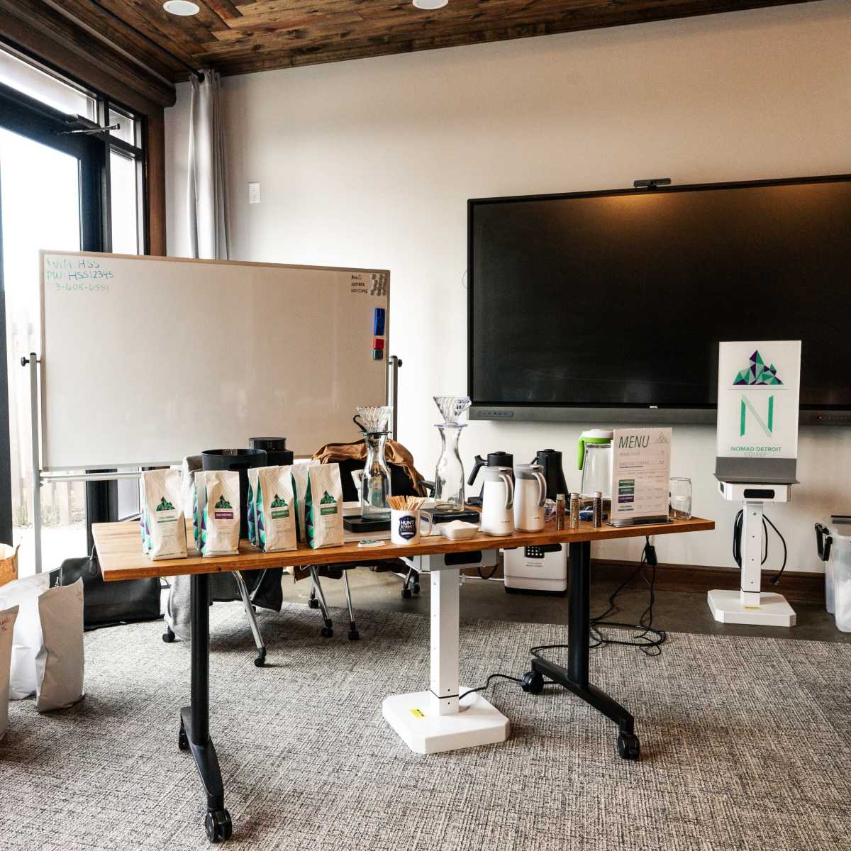 A conference room is set up with a large table displaying various coffee equipment, a whiteboard, and a television screen on the wall.