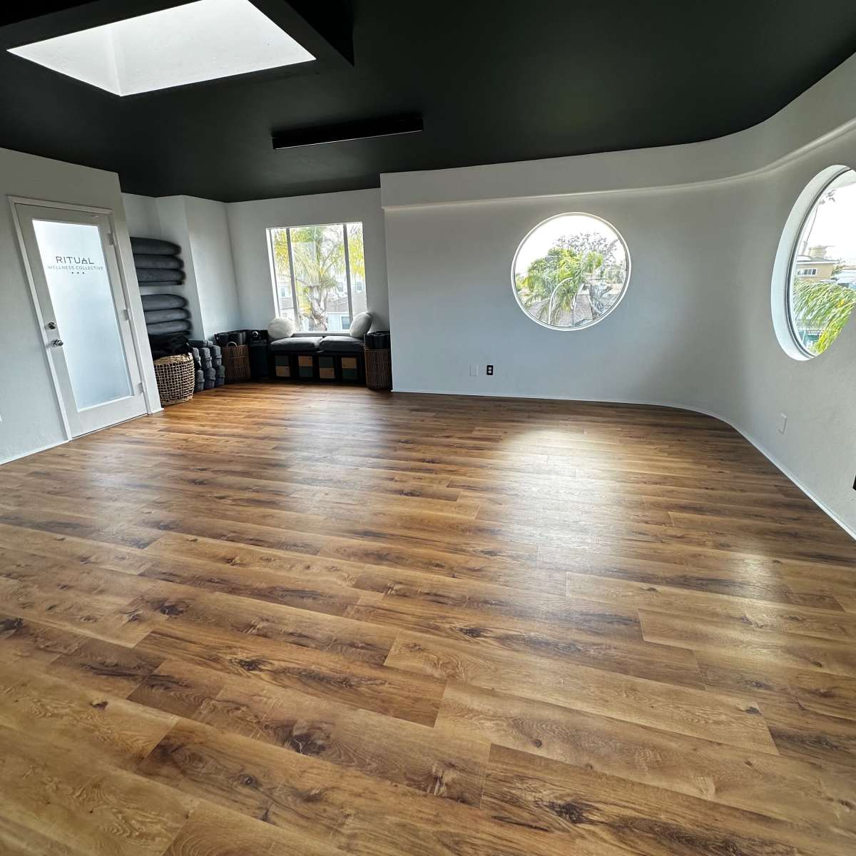 A spacious room with wooden flooring, a black ceiling, and circular windows providing natural light.