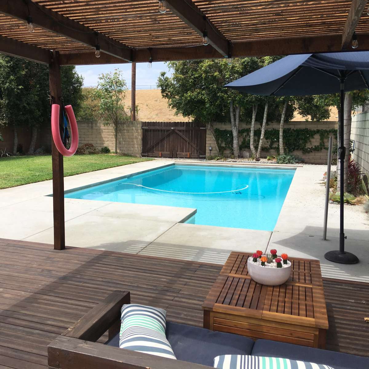 A well-maintained backyard features a swimming pool surrounded by a patio, lounge furniture, and a covered area with a hanging ring.