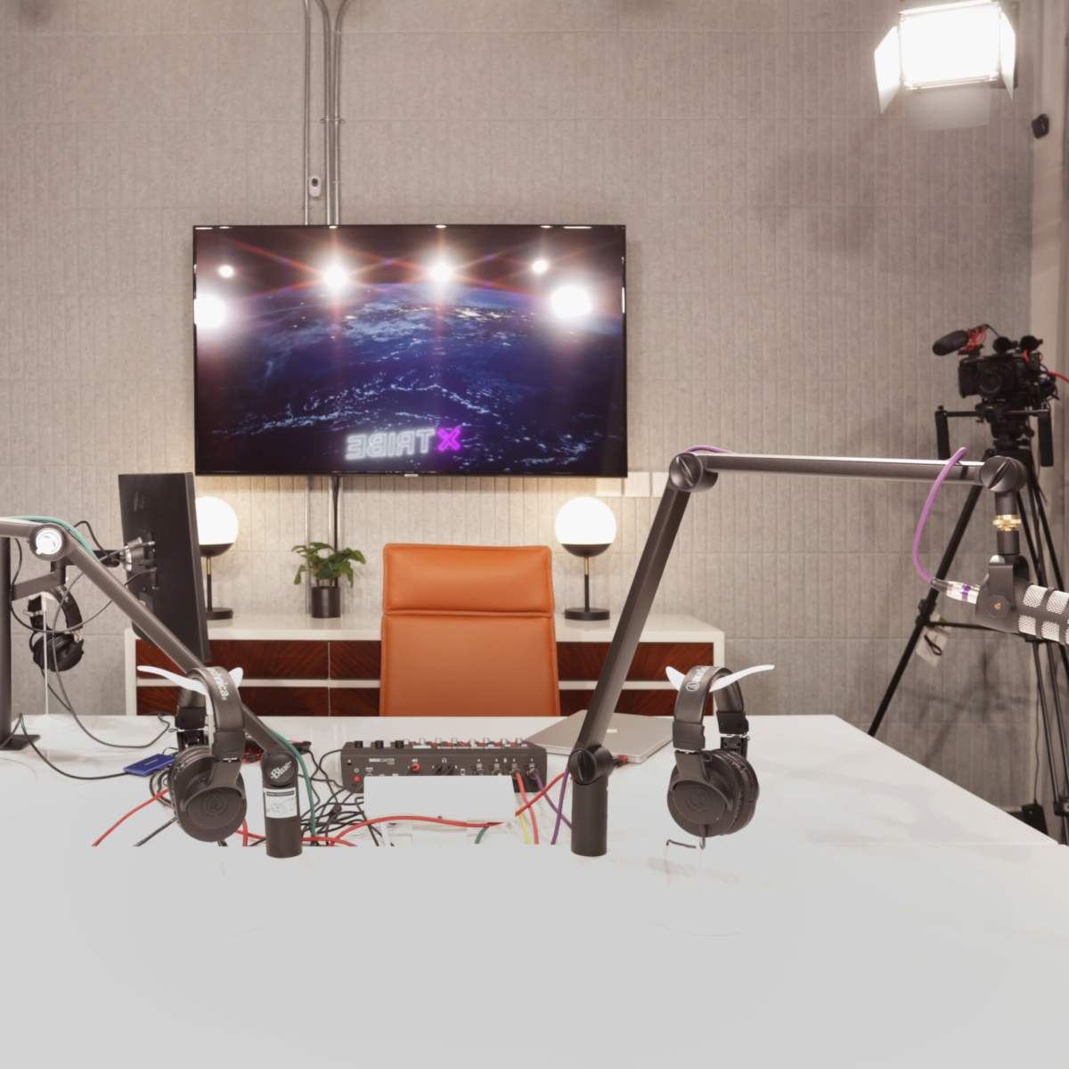 A podcast recording studio features two microphone setups, a central table with audio equipment, and a TV screen displaying a graphic.