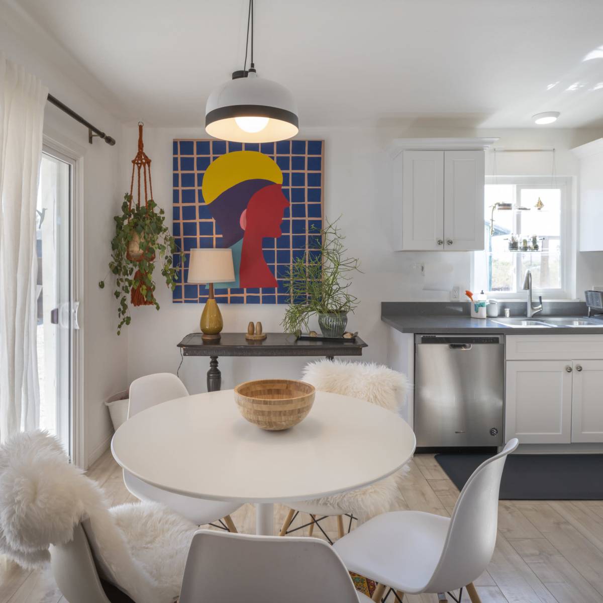 A bright kitchen features a round dining table with a wooden bowl, surrounded by white chairs, and is adorned with a colorful abstract wall art and plants.