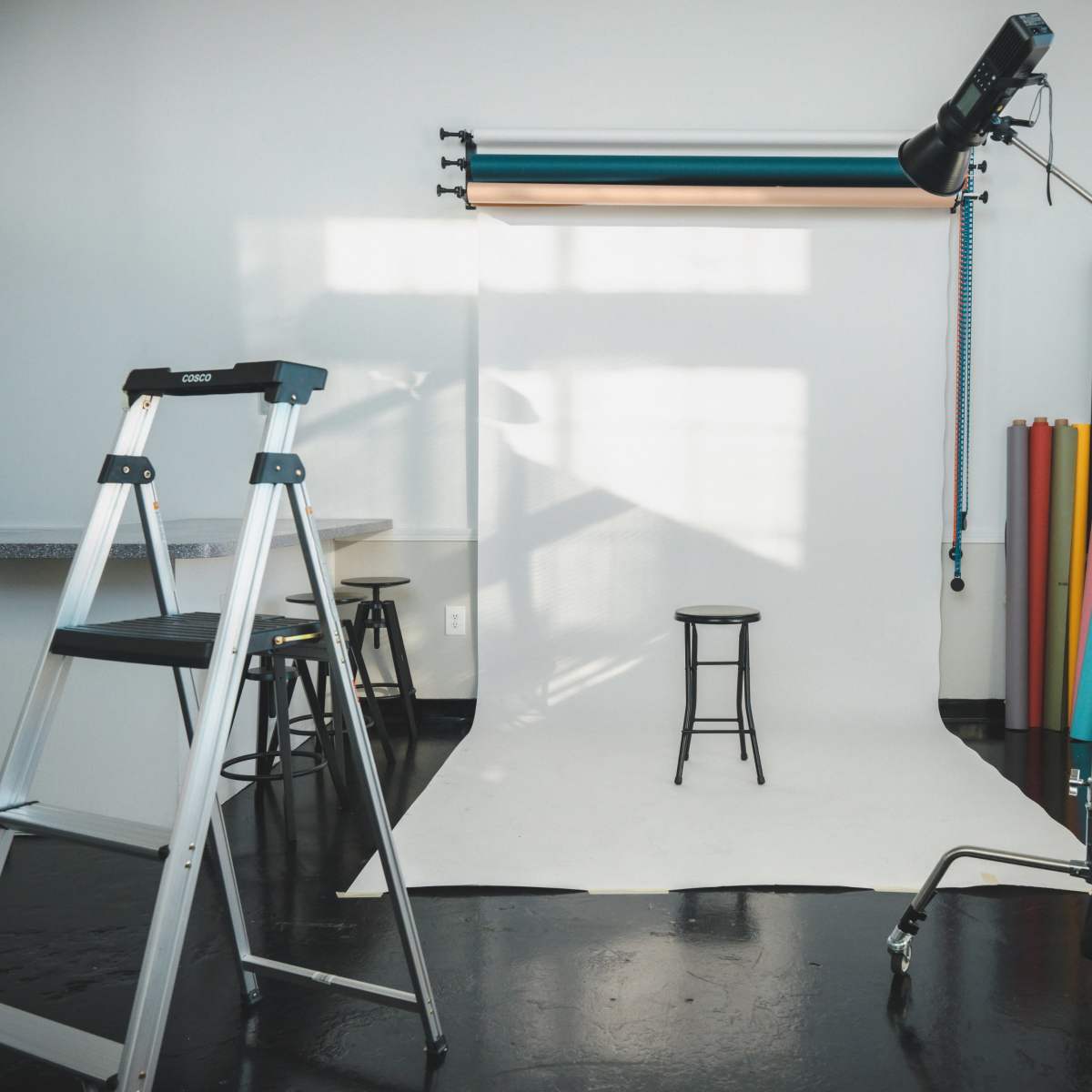 A photography studio with a white backdrop, a stool in the center, and various lighting equipment and props arranged around a ladder.
