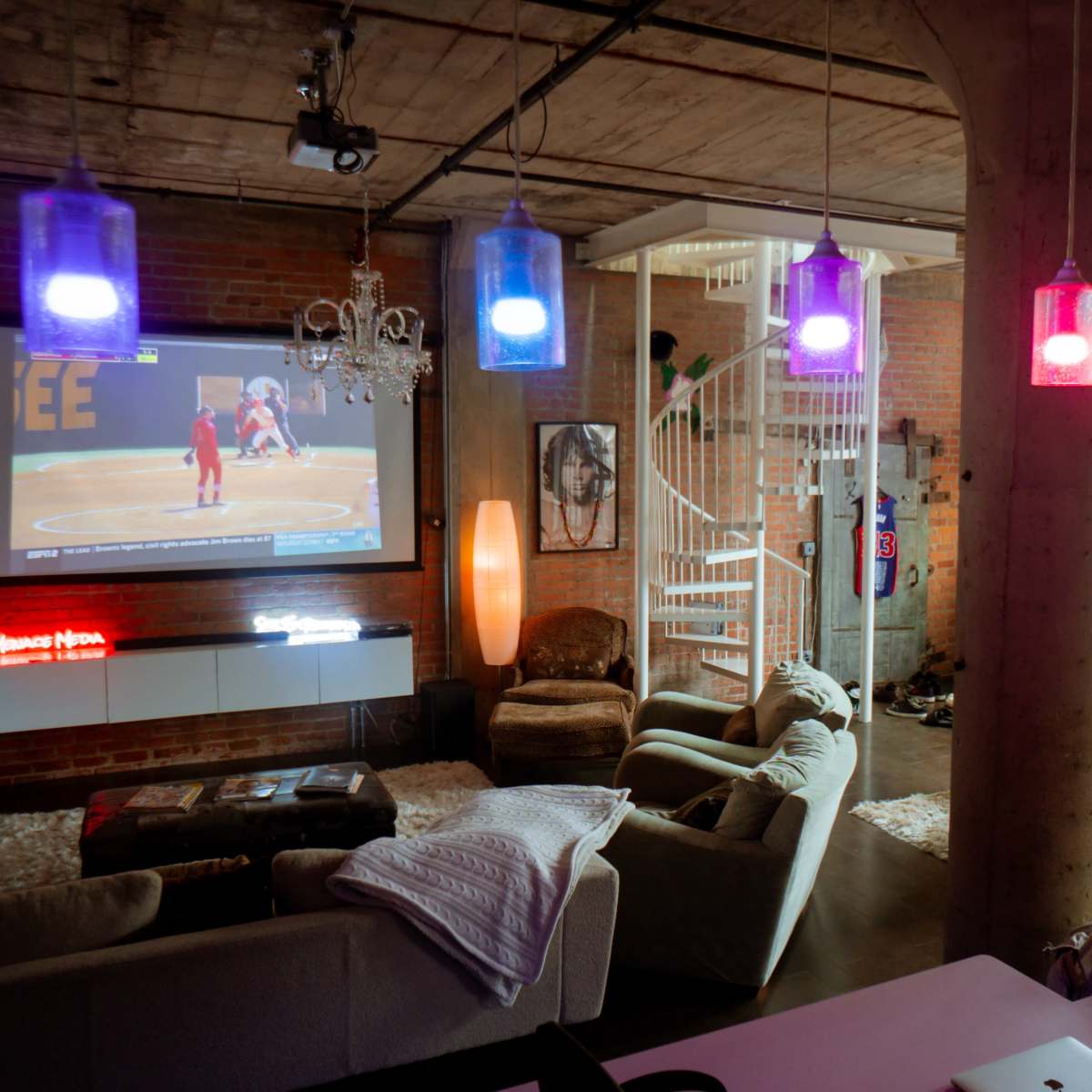 A modern loft-style living area featuring a large screen displaying a sports game, colorful pendant lights, and a mix of furniture arrangements including a dining table and seating areas.