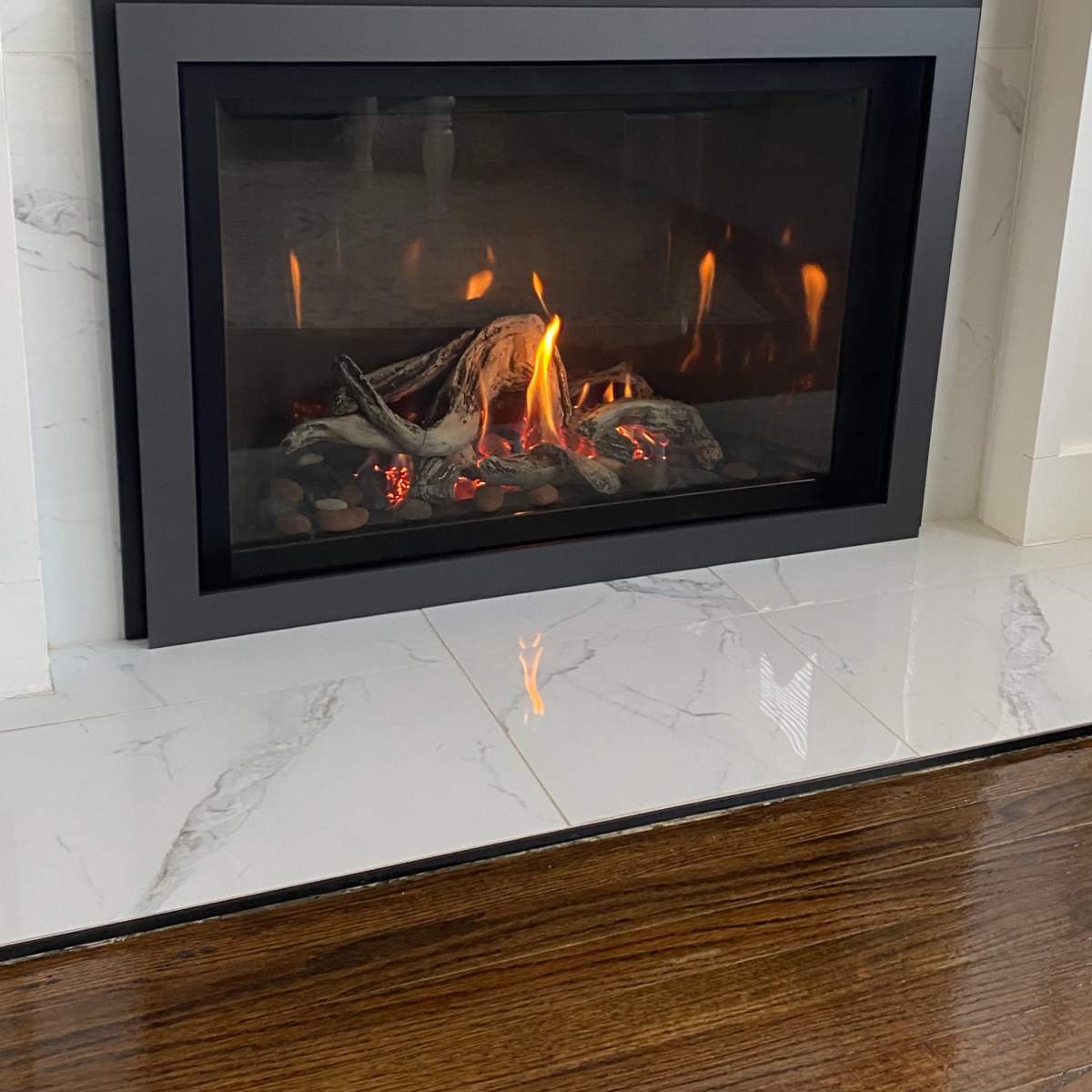 A modern gas fireplace with a glass front is set into a white marble wall, displaying flames and glowing logs on a bed of stones.