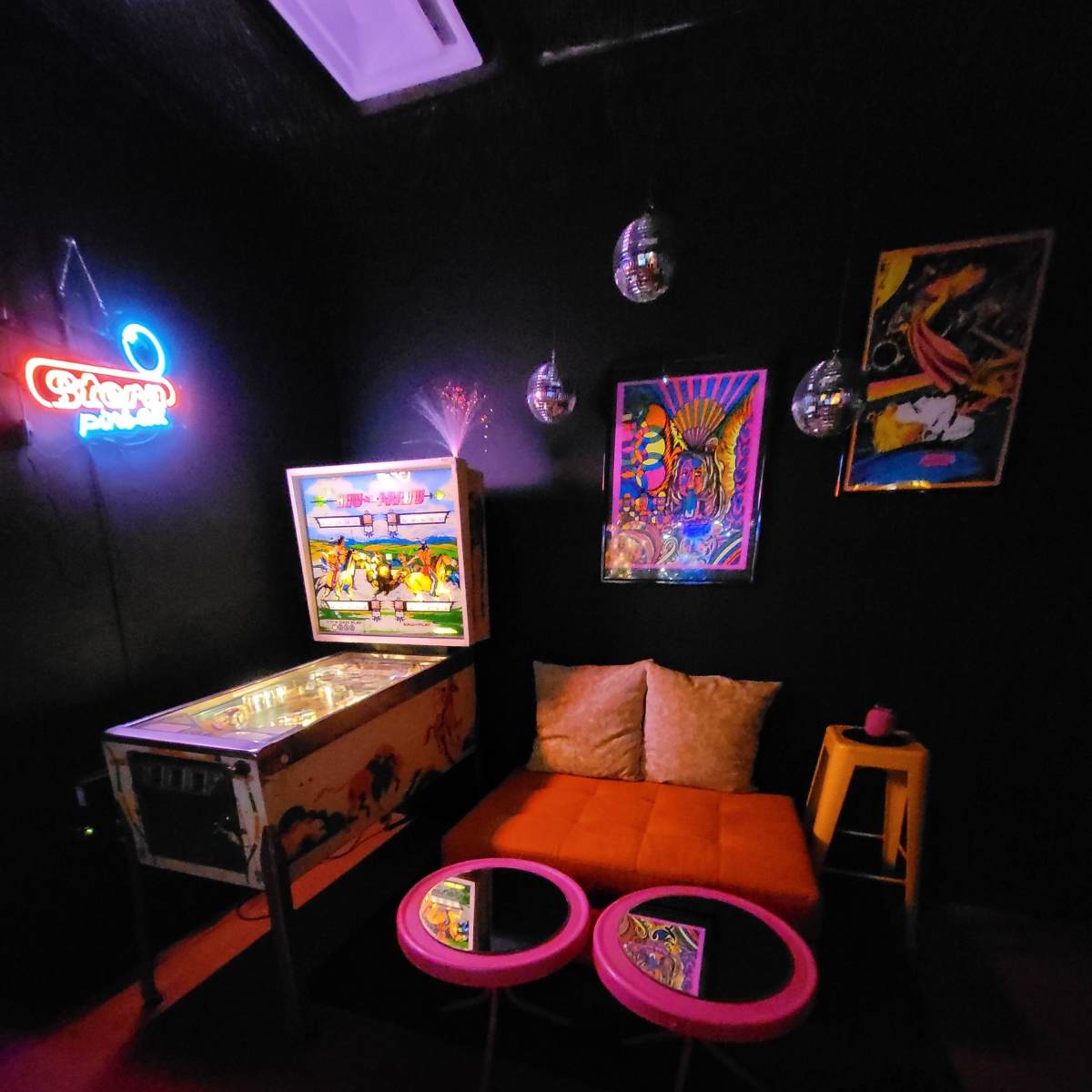 A cozy lounge area with an orange couch, two pink coffee tables, a vintage pinball machine, and colorful artwork on a black wall.
