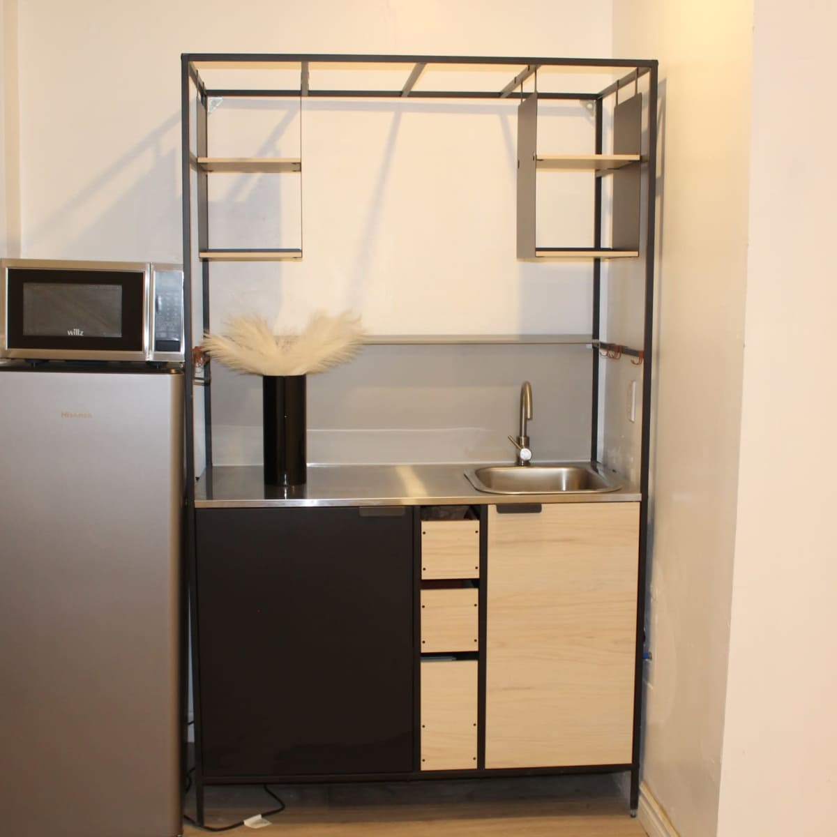 A modern kitchen setup featuring a stainless steel refrigerator, a microwave on a shelf, and a compact sink with cabinetry below.