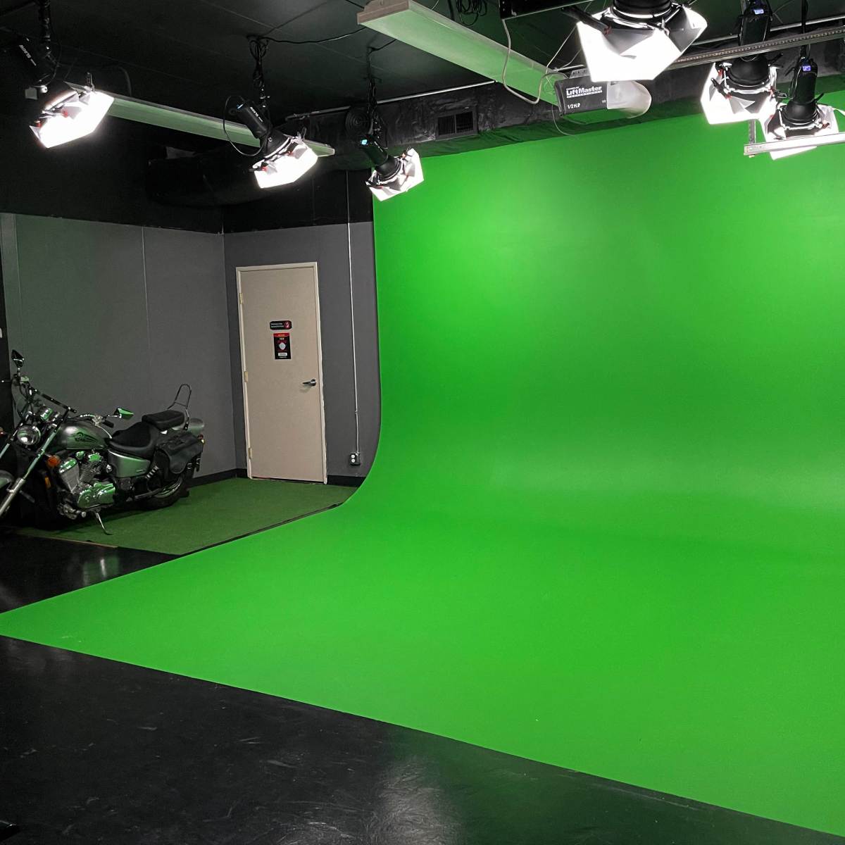 A studio space featuring a large green screen background, equipped with lighting fixtures and a motorcycle positioned on the floor.