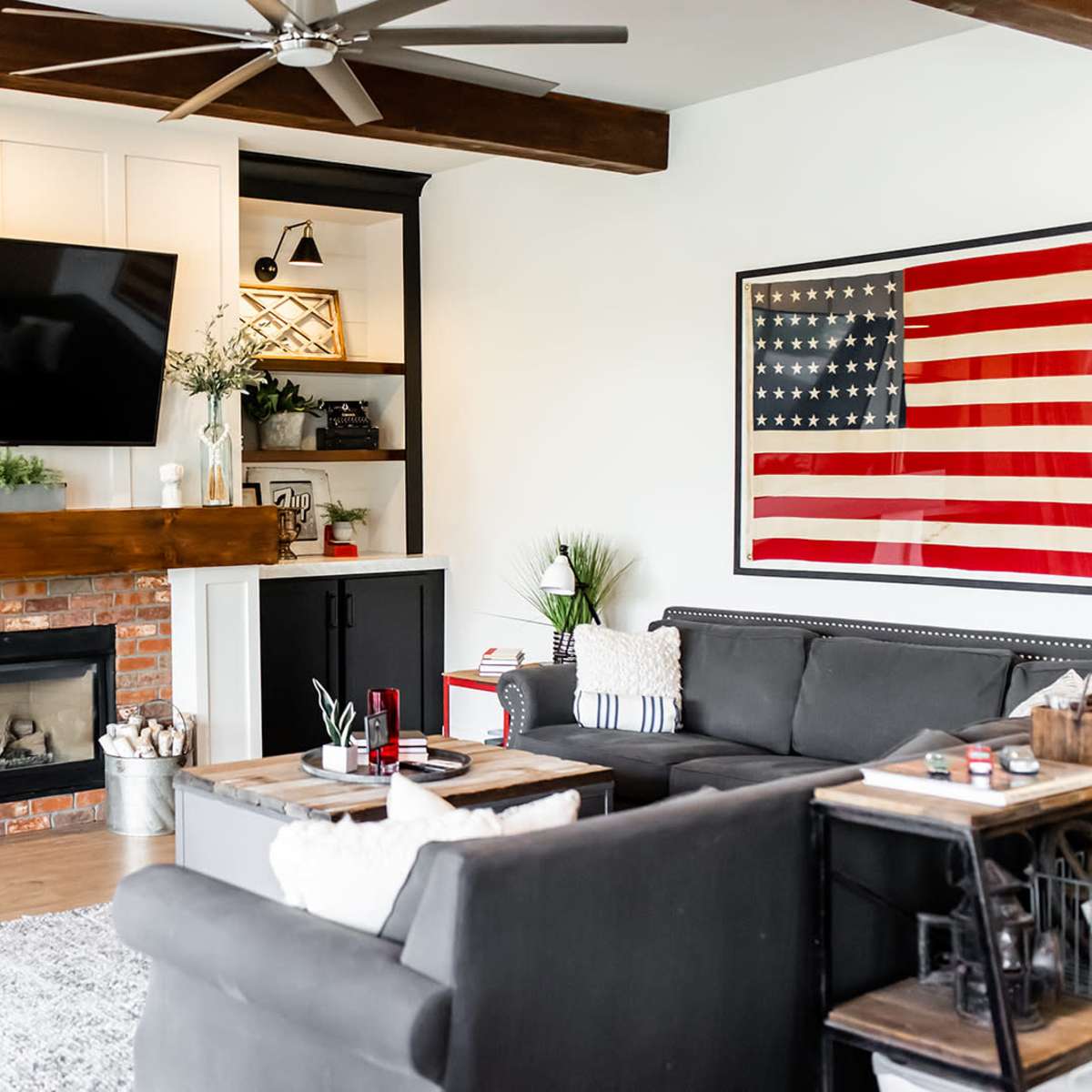 A modern living room features a large television above a brick fireplace, flanked by built-in shelves, with a rustic wooden mantel and an American flag artwork on the wall.