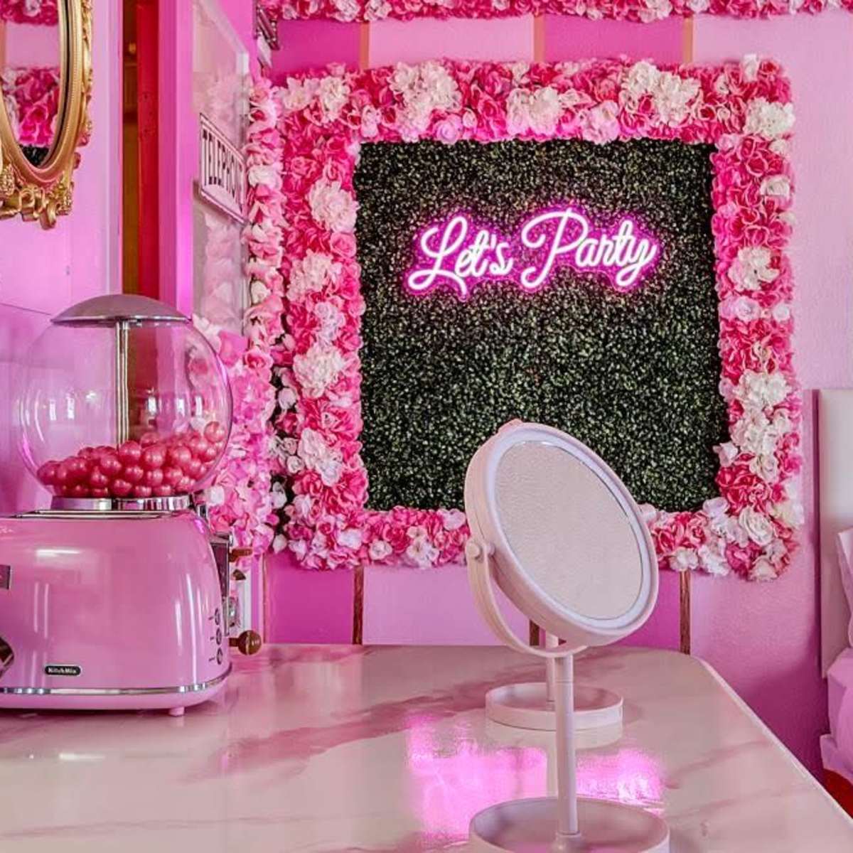 A pink-themed room featuring a neon sign that reads "Let's Party," surrounded by floral decorations and a vintage-style gumball machine.