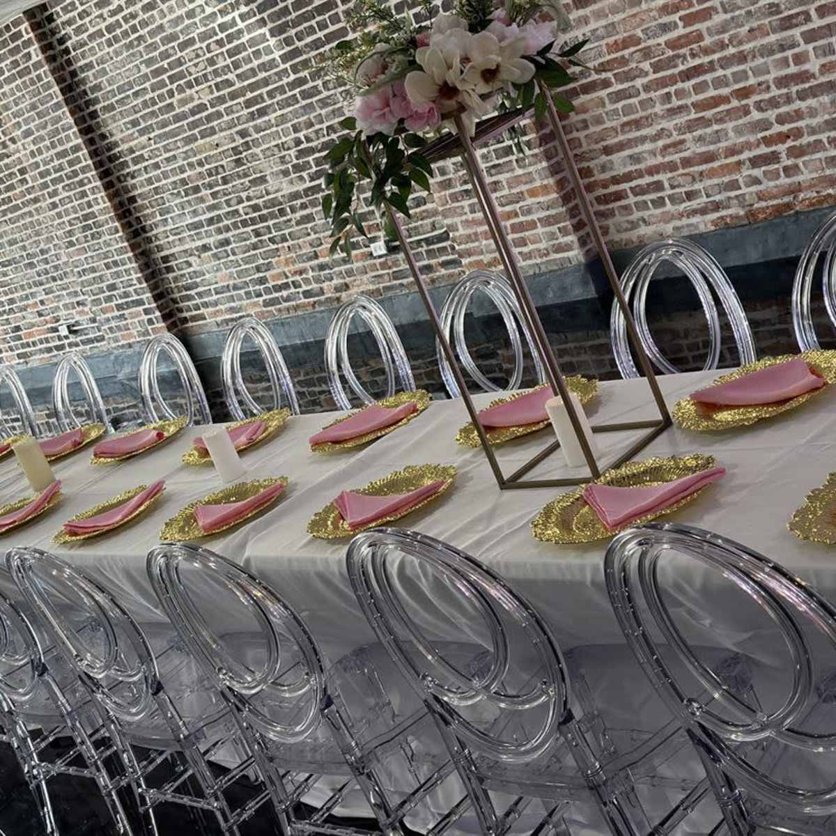 A long rectangular dining table is set with pink napkins, decorative gold chargers, and clear acrylic chairs in a room with exposed brick walls.