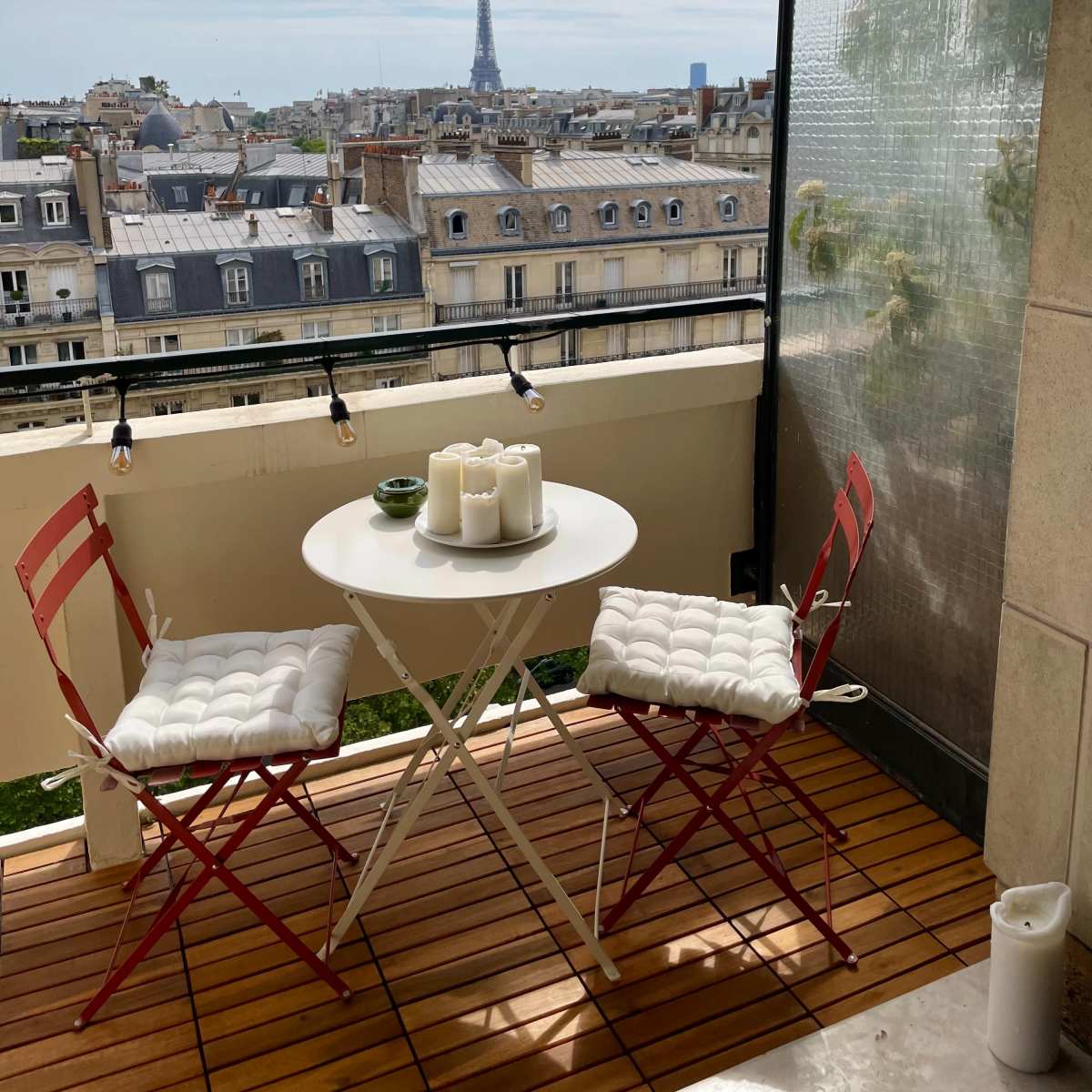 A small balcony features a round white table with two red chairs, overlooking a cityscape that includes the Eiffel Tower in the distance.