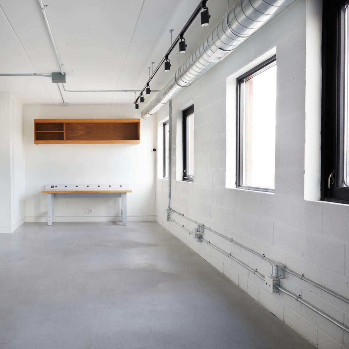 A spacious, empty room with concrete flooring, white walls, windows, and a wooden shelf mounted above a long table.