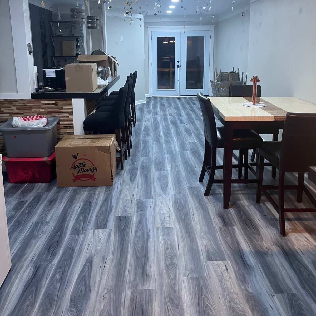 A partially furnished interior space with a dining table and chairs, a counter with bar stools, and boxes stacked along the walls, all beneath a ceiling decorated with hanging stars.