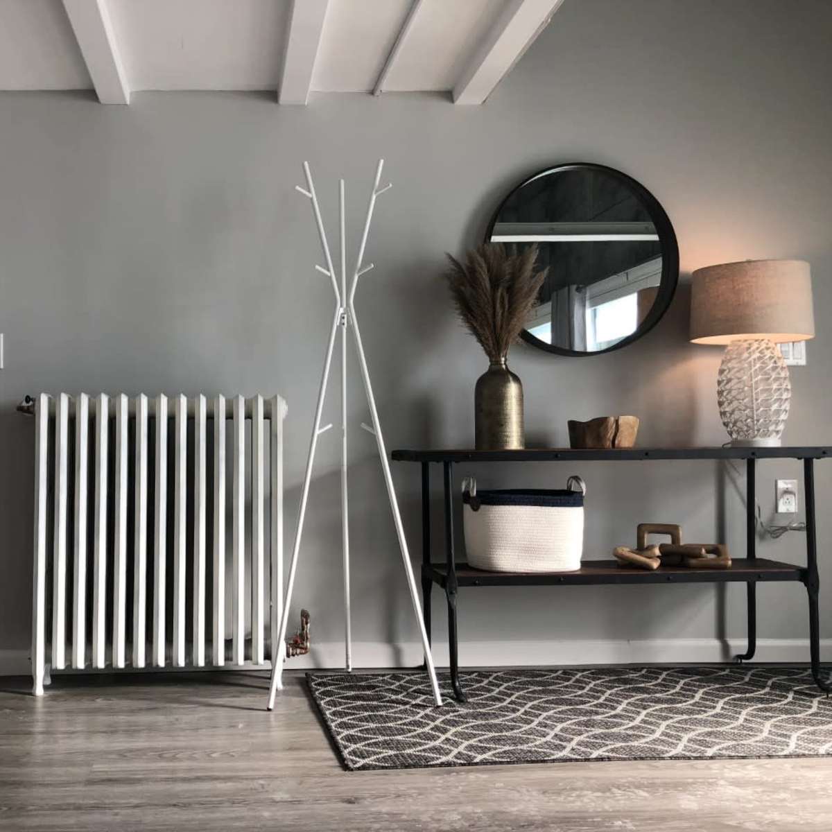 A minimalist interior with a white radiator, a coat stand, a round mirror, a lamp, and a table displaying a decorative basket and vase.