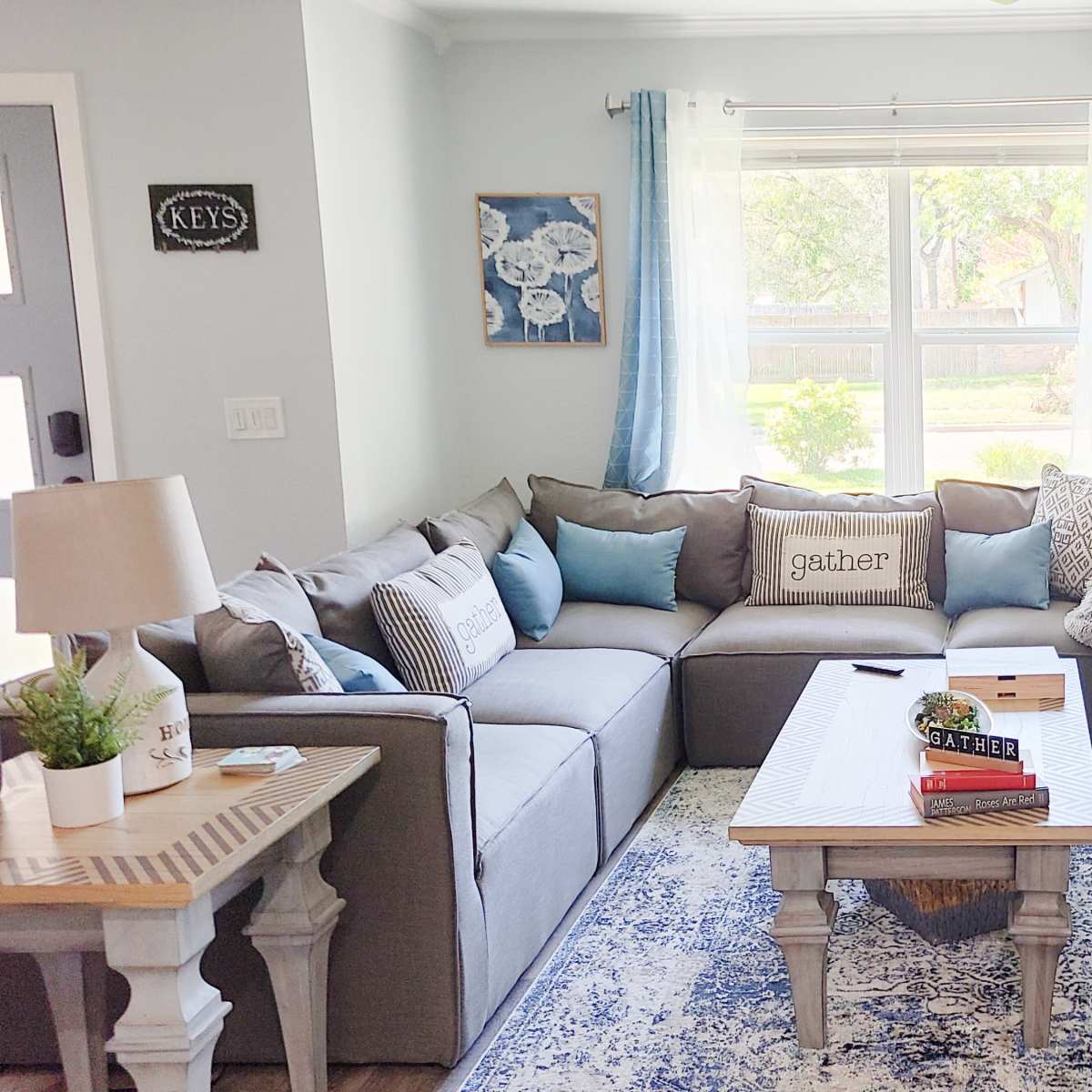 The living room features a gray sectional sofa adorned with blue and white cushions, a wooden coffee table, and a large TV on a console, all arranged around a window that lets in natural light.
